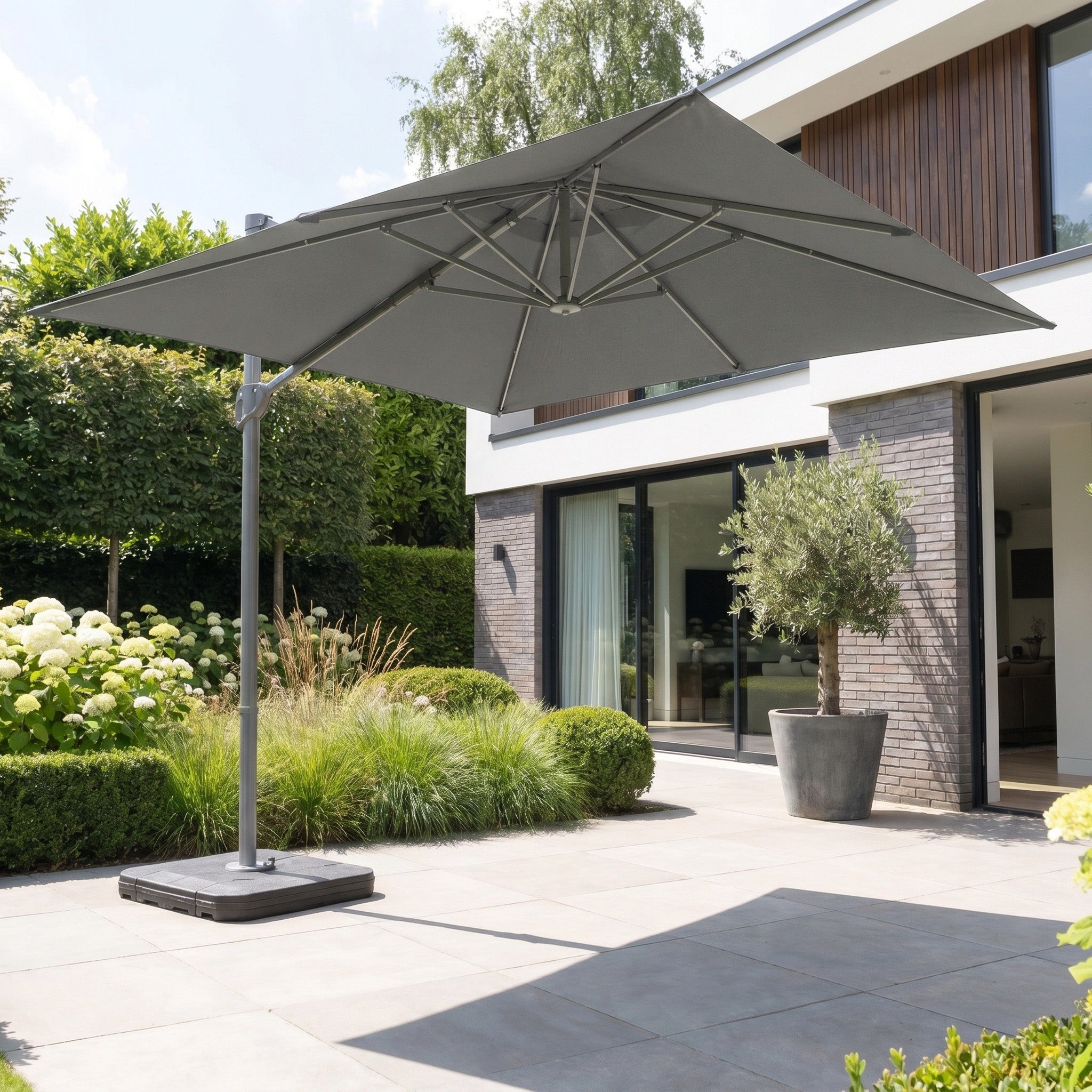 The Hera 2.7m Square Cantilever Parasol with LED Lighting in Charcoal stands open on a modern terrace next to glass doors and lush plants.