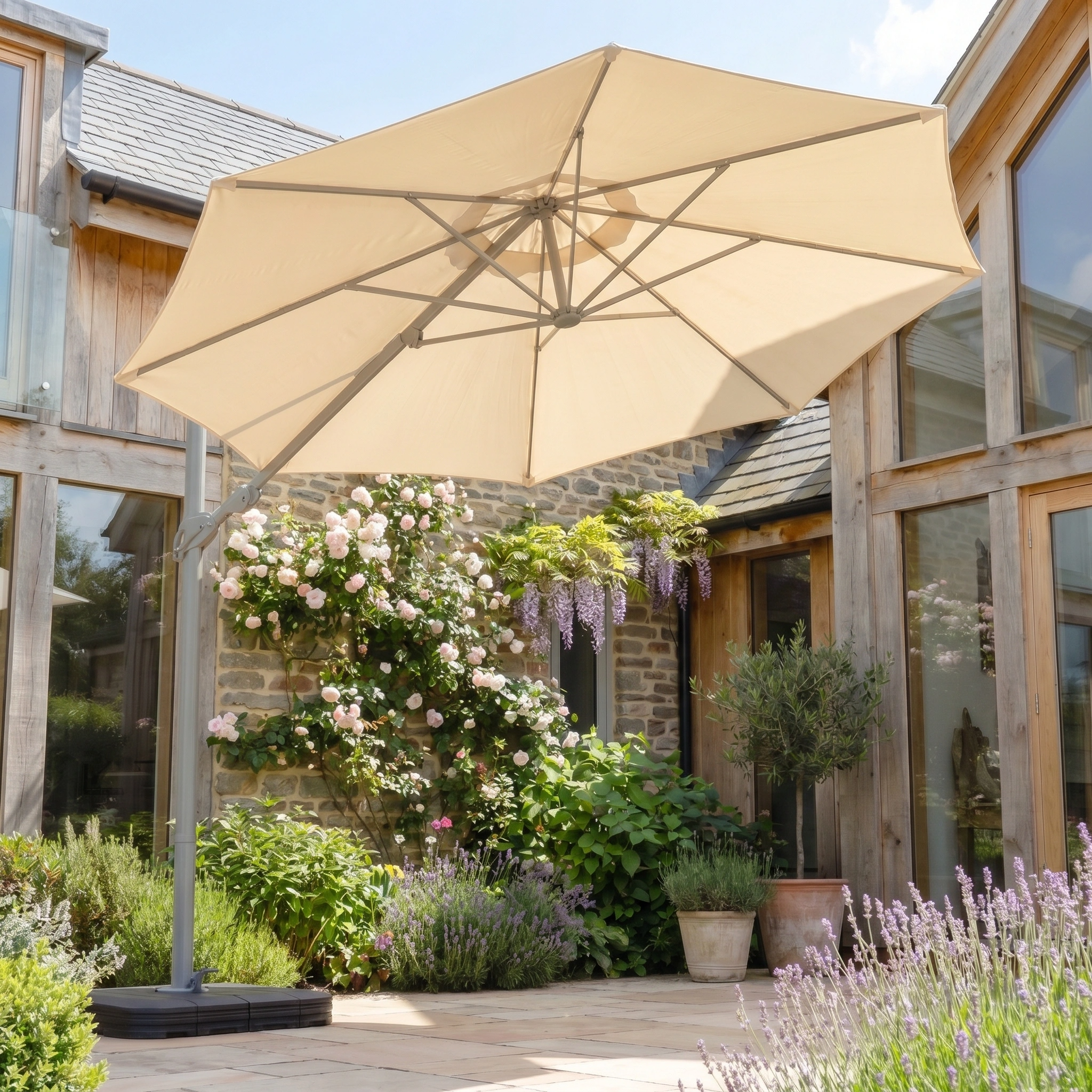 The Hera 3m Round Cantilever Parasol with LED Lighting in Beige stands in a sunny garden courtyard, surrounded by flowers and plants near a charming stone house.