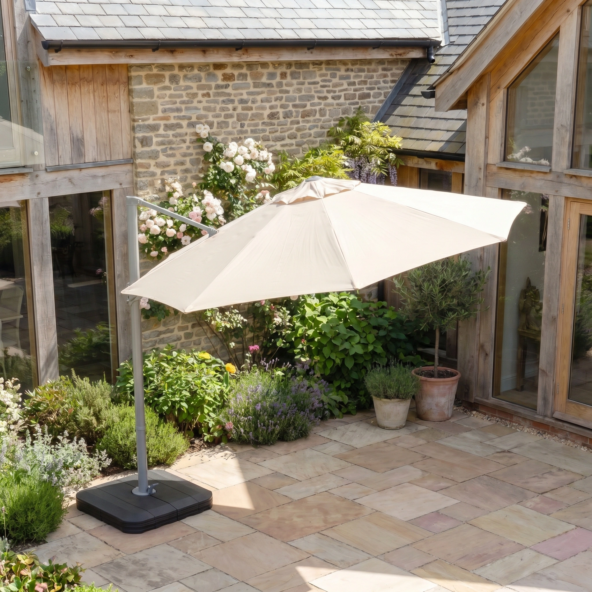 The Hera 3m Round Cantilever Parasol with LED Lighting in Beige provides shade for a stone courtyard, set amidst lush plants and wooden buildings.