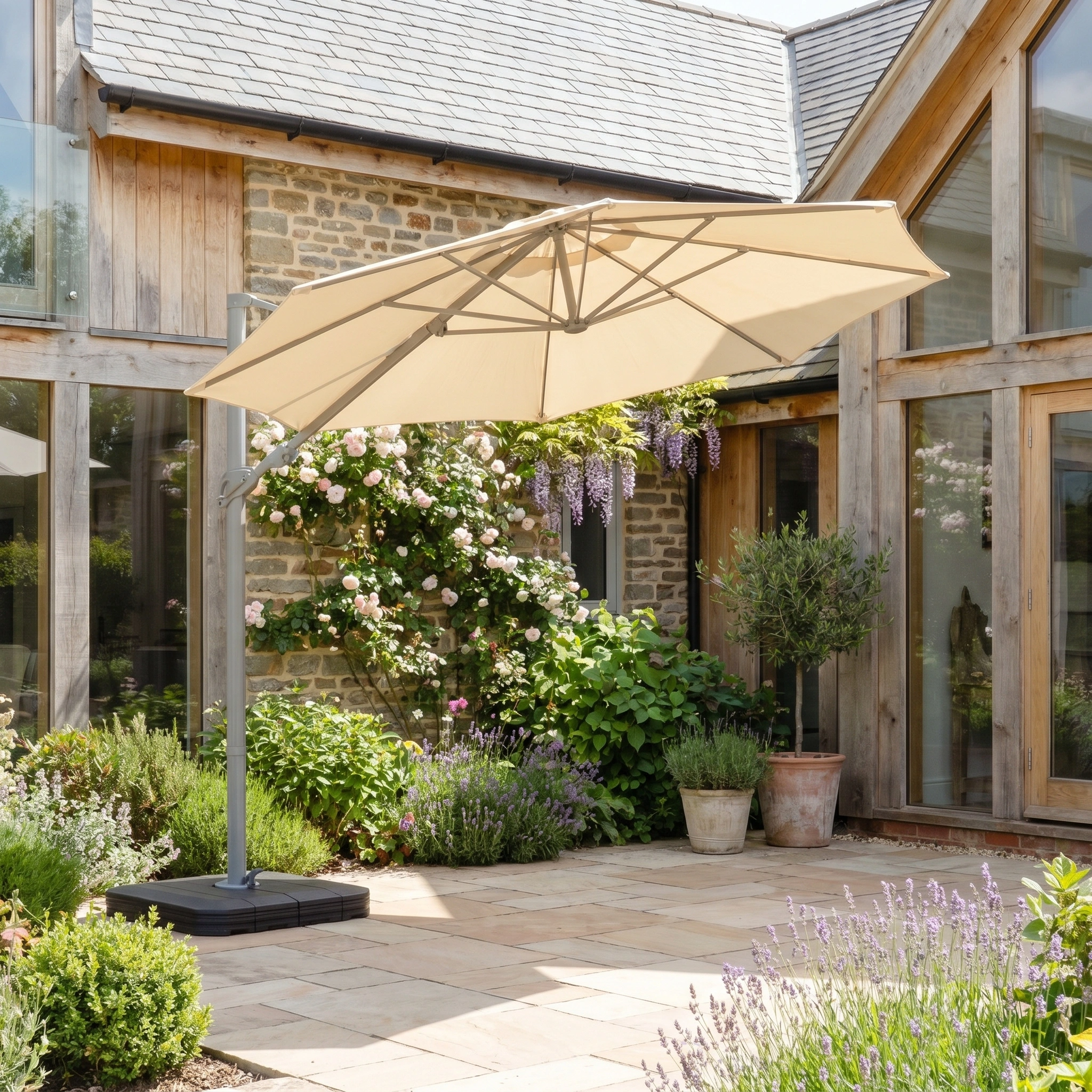 The Hera 3m Round Cantilever Parasol with LED Lighting in Beige provides UV50+ shade for a stone terrace with potted plants and blooming flowers beside a modern house.