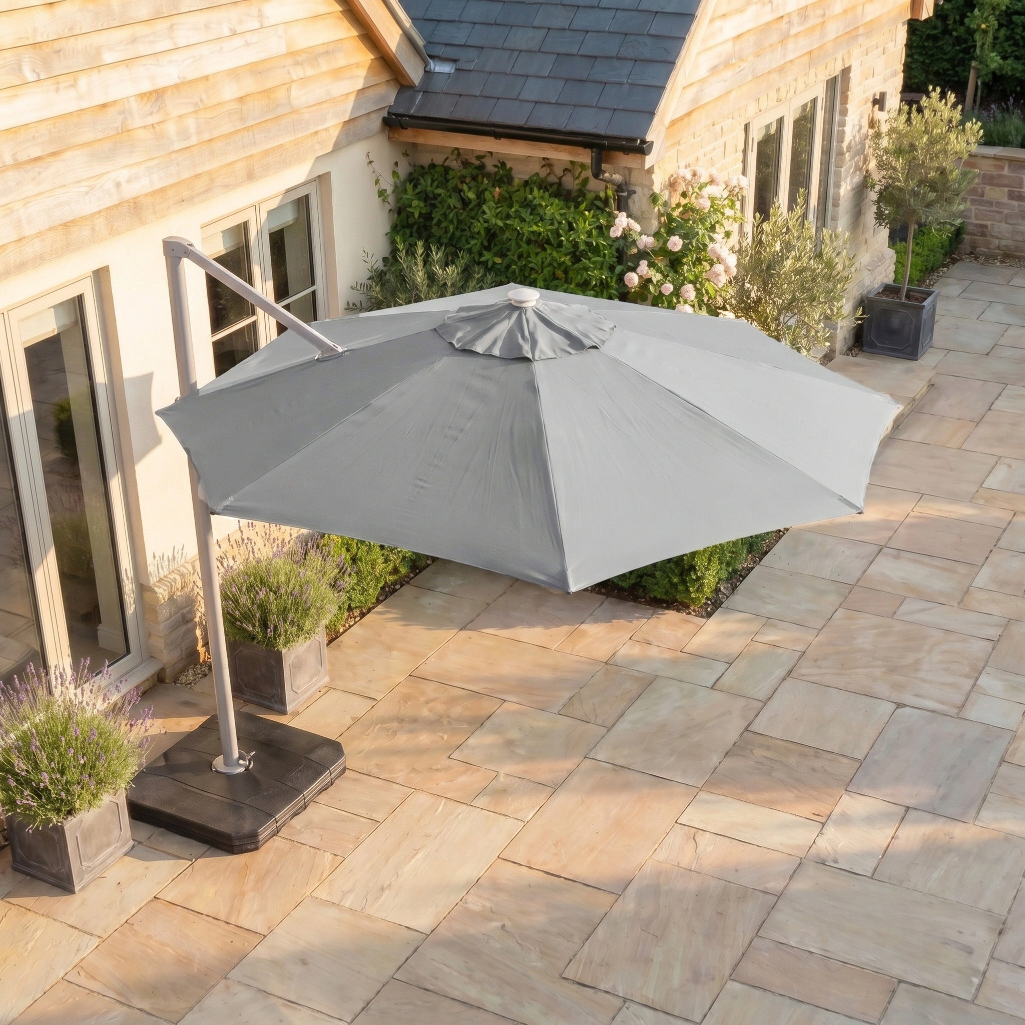 The Hera 3m Round Cantilever Parasol with LED Lighting in Grey stands on a stone-tiled terrace next to a house, surrounded by potted plants and shrubs.