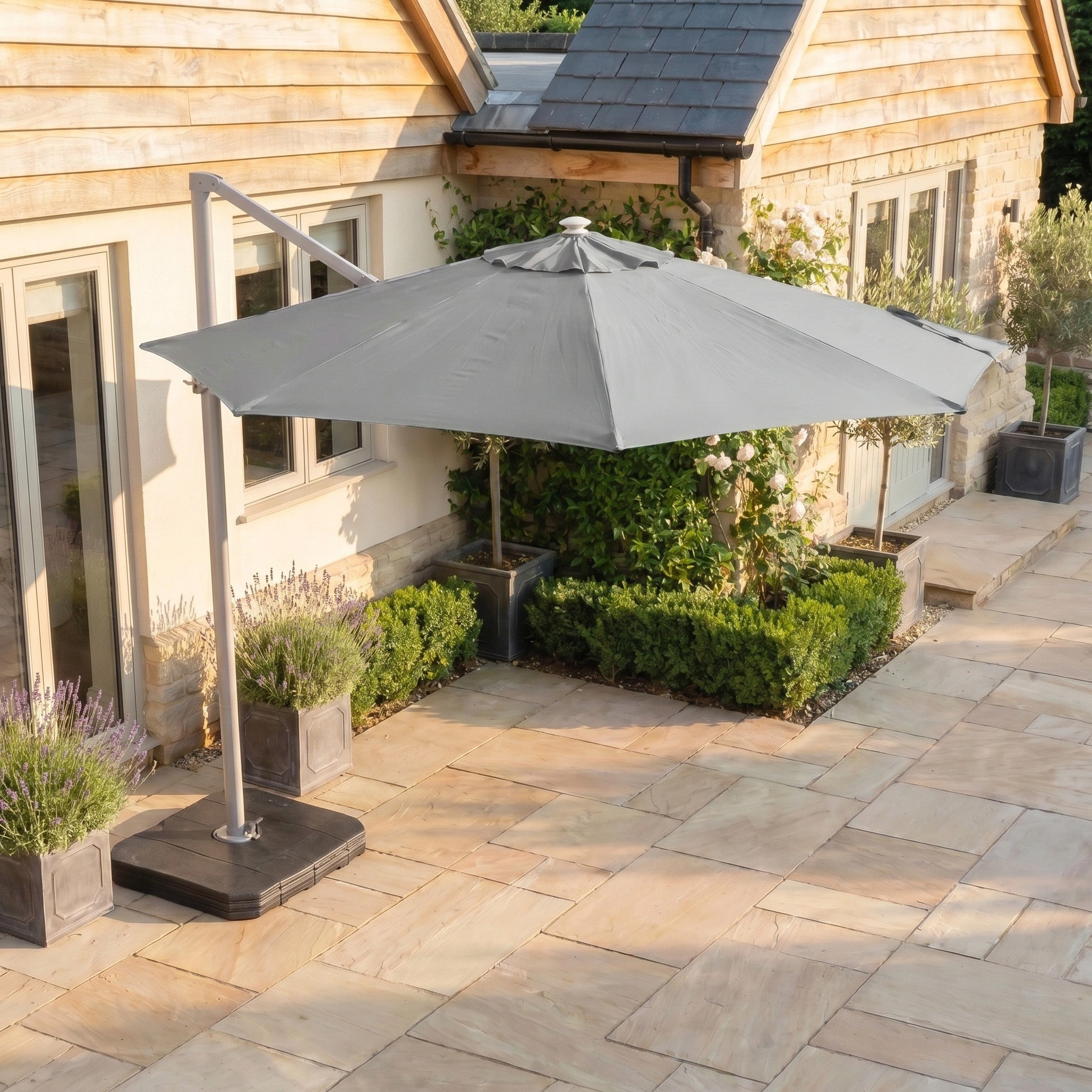 The Hera 3m Round Cantilever Parasol with LED Lighting in Grey, featuring a UV50+ canopy, shades a stone patio next to a house decorated with potted plants and shrubs.