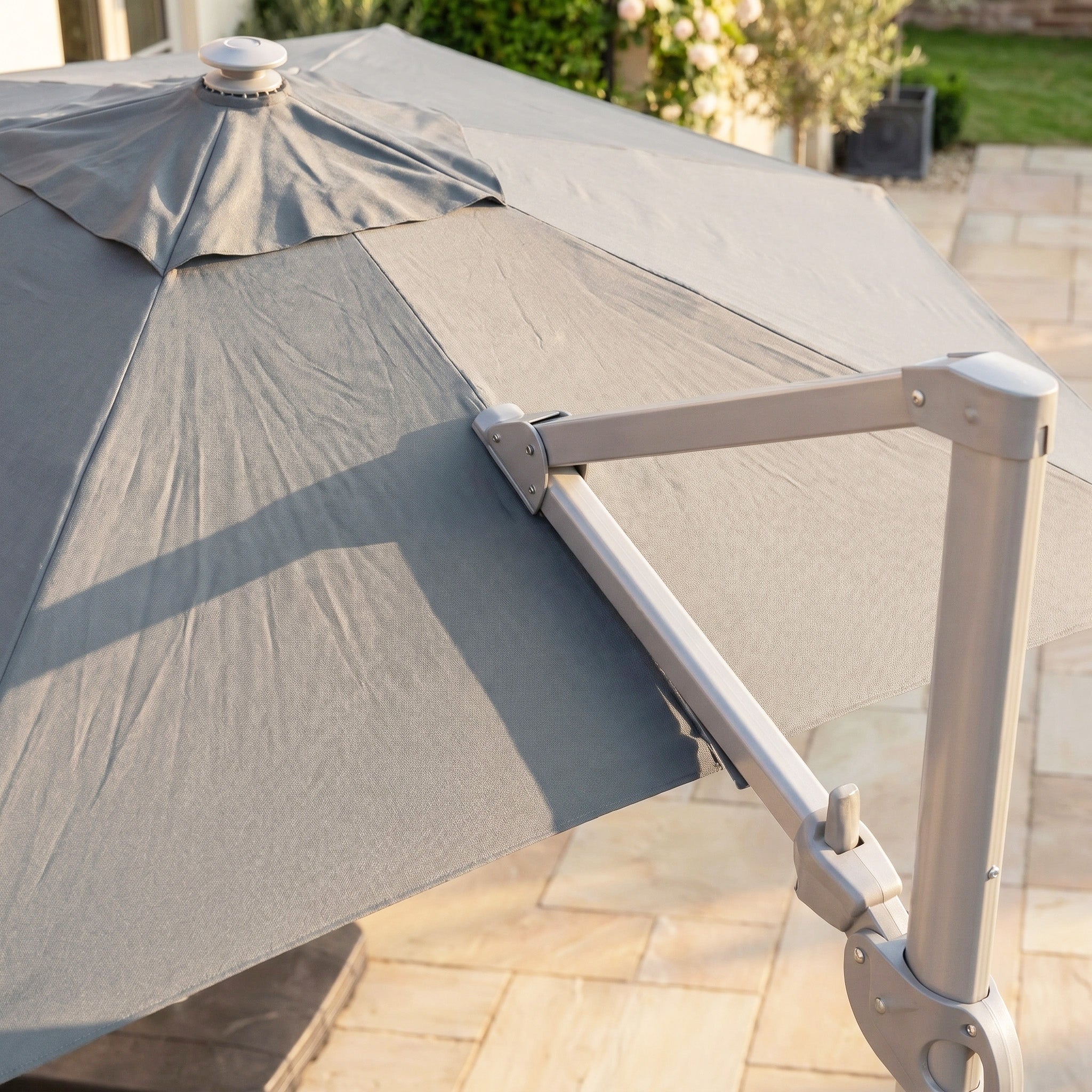 The Hera 3m Round Cantilever Parasol with LED Lighting in Grey stands on a stone-tiled patio, its metal frame and UV50+ canopy basking in sunlight.