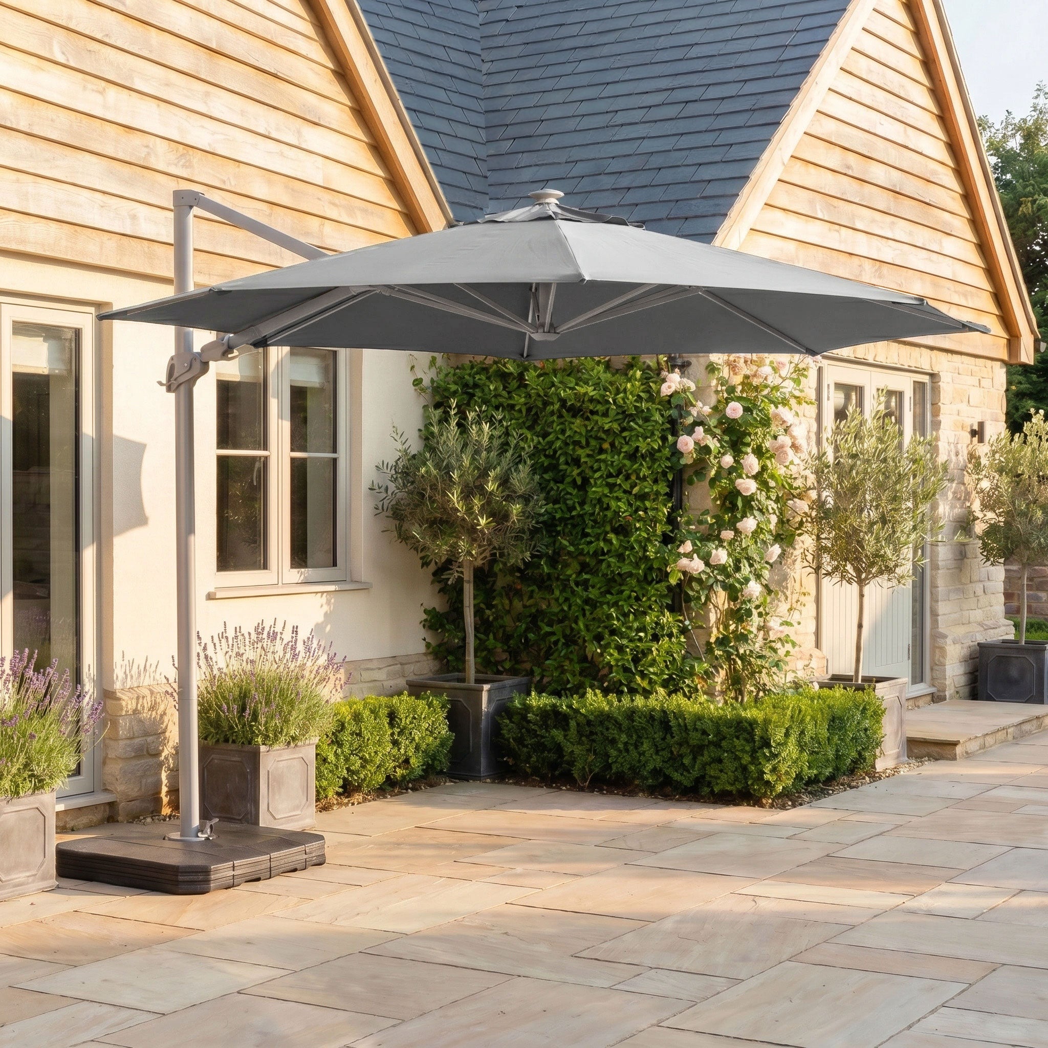 The Hera 3m Round Cantilever Parasol with LED Lighting in Grey provides UV50+ shade for a stone terrace beside a house with wooden siding and potted plants.