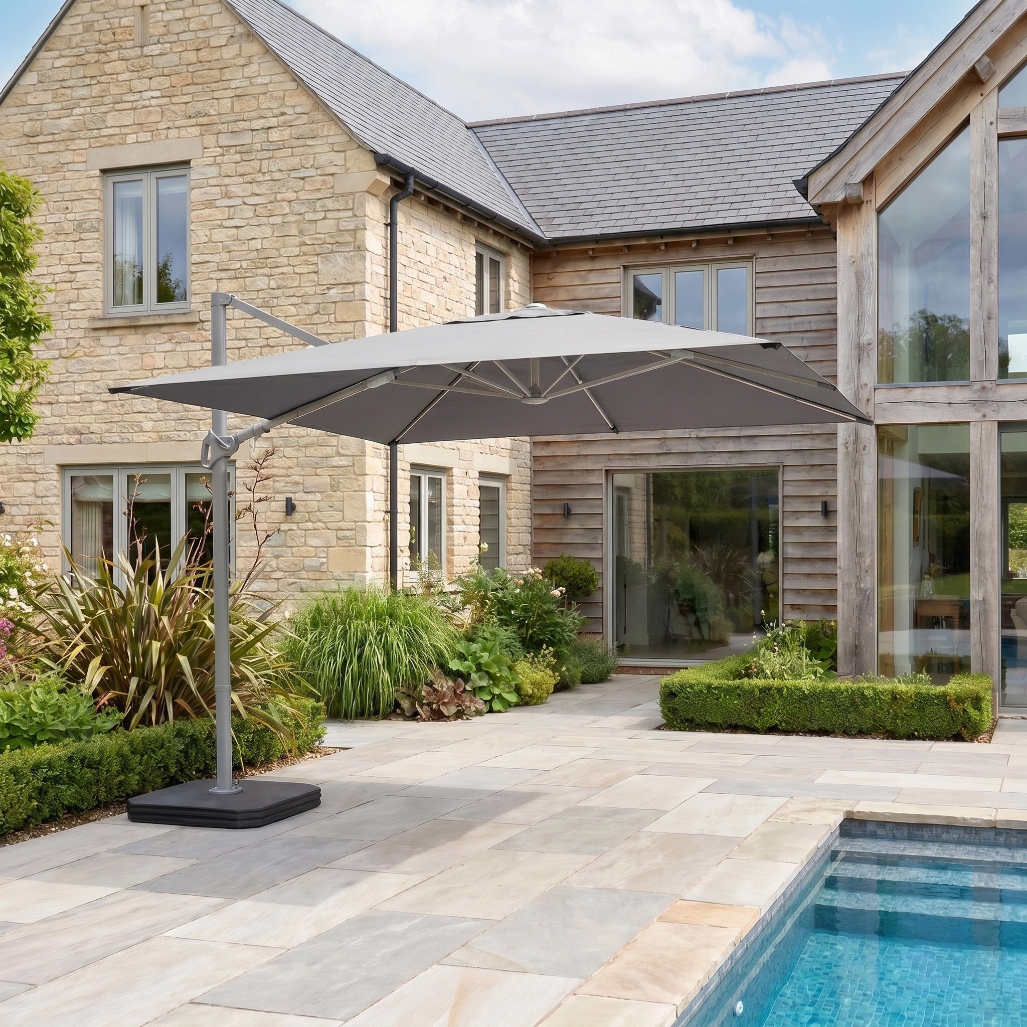The Hera 3m x 2m Rectangular Cantilever Parasol with LED Lighting in Grey provides shade for a stone-paved backyard near a pool, adjacent to a modern stone house.