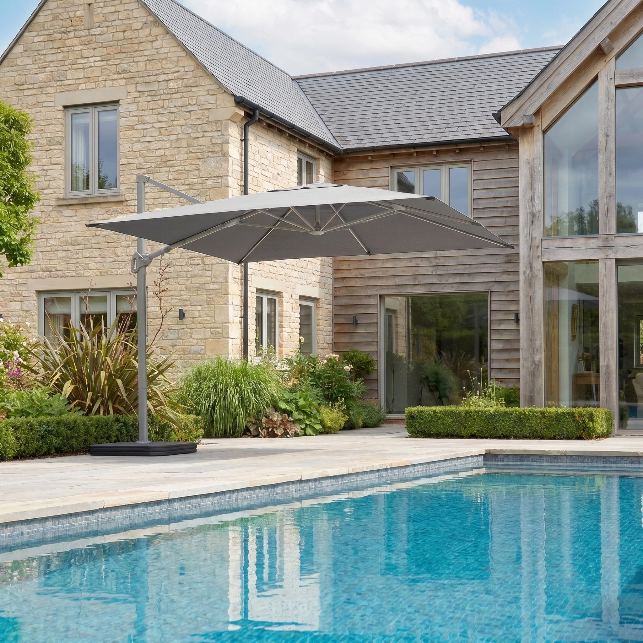 Modern house with stone and wood exterior, poolside patio, lush green landscaping, and a Hera 3m x 2m Rectangular Cantilever Parasol with LED Lighting in Grey.
