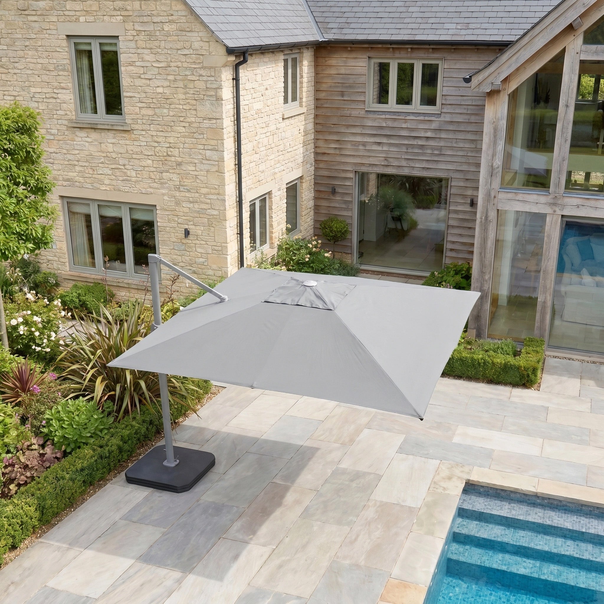 The Hera 3m x 2m Rectangular Cantilever Parasol with LED Lighting in Grey shades the stone-tiled poolside in a modern garden, with a stylish house visible in the background.