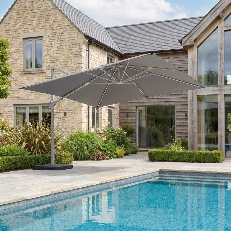 The Hera 3m x 2m Rectangular Cantilever Parasol with LED Lighting in Charcoal provides shade by the pool outside a stone and wood house with gardens.