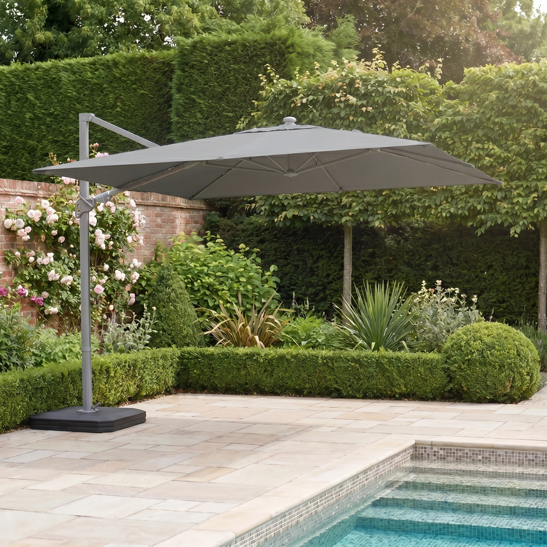 A Hera 3m x 2m Rectangular Cantilever Parasol with LED lighting in Charcoal and a UV50+ canopy stands beside a pool in a landscaped garden with hedges and flowering plants.