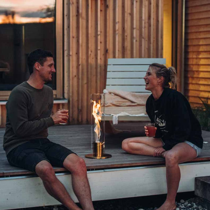 Two people are seated on a wooden deck, enjoying drinks and talking beside a modern, glass-enclosed patio fire pit. They are dressed casually and appear relaxed.