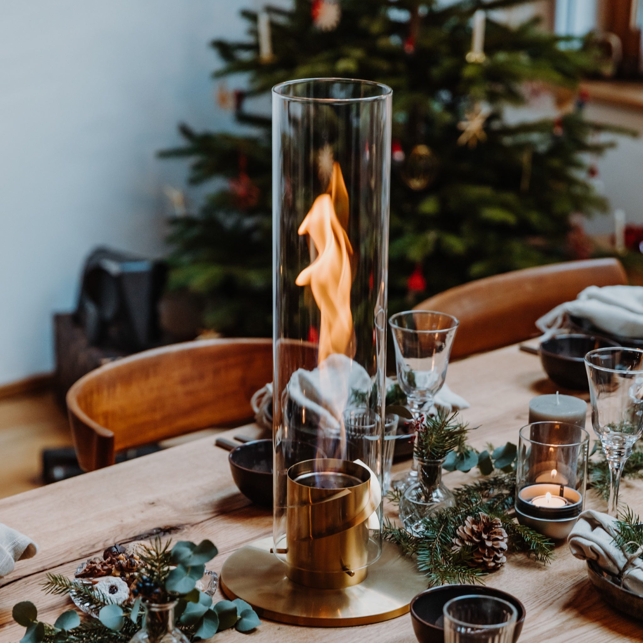 A dining table with a modern glass and metal candle holder, festive decorations, and a Höfats SPIN 90 Table Top Fire in Gold centerpiece in the background.
