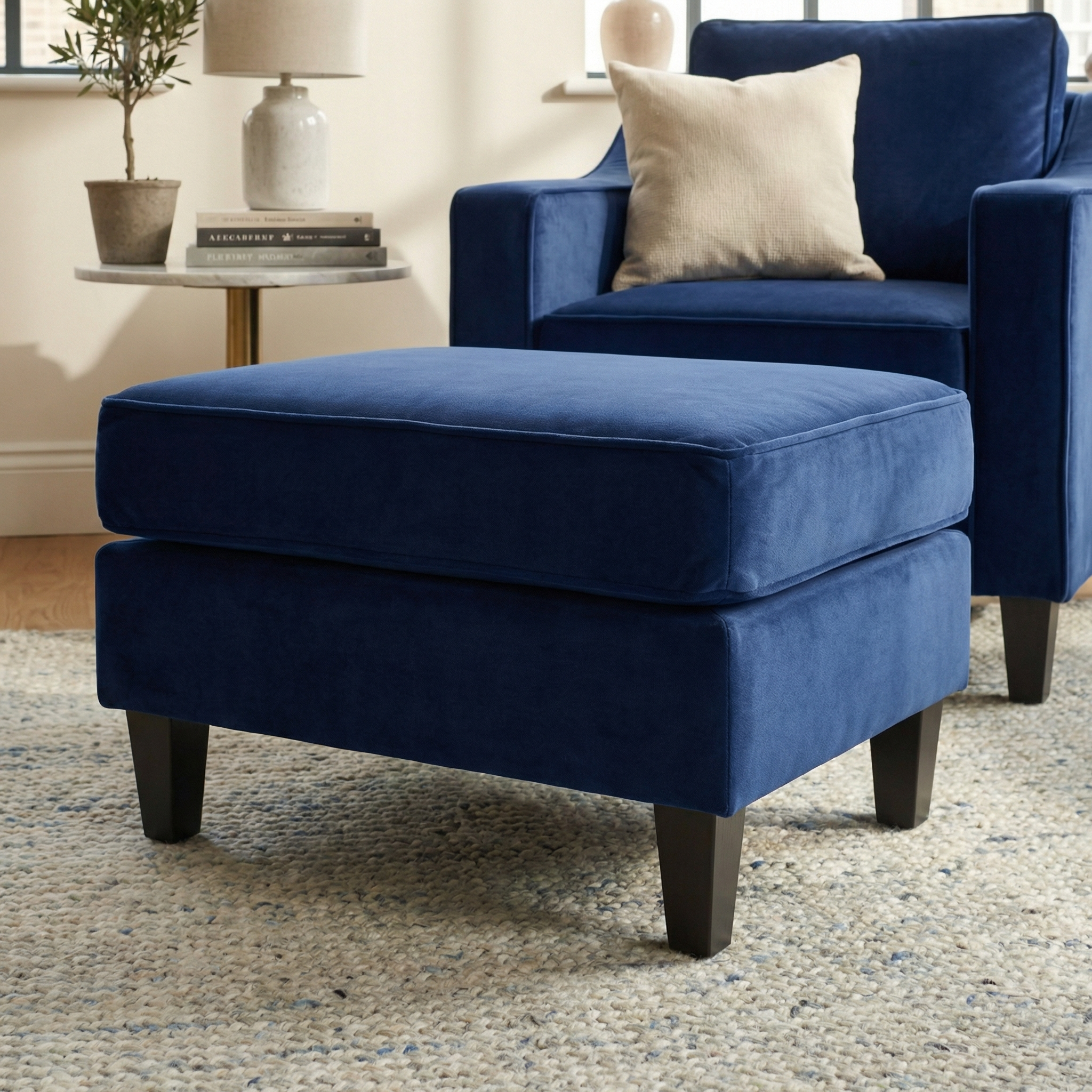 The Wiltshire Velvet Footstool in Royal Blue with dark wood legs sits before a matching armchair, adding a stylish touch to the neutral decor.