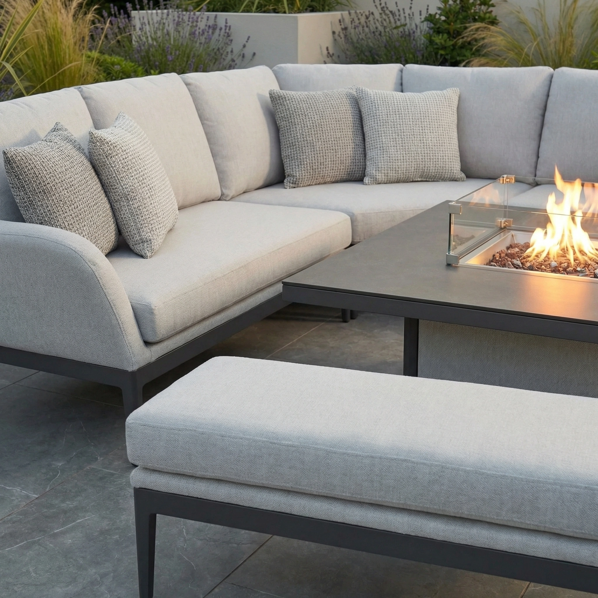 Modern patio featuring the Luna Deluxe Outdoor Fabric Square Corner Dining Set with Rising Firepit Table in Oyster Grey, set on a stone floor and surrounded by greenery.