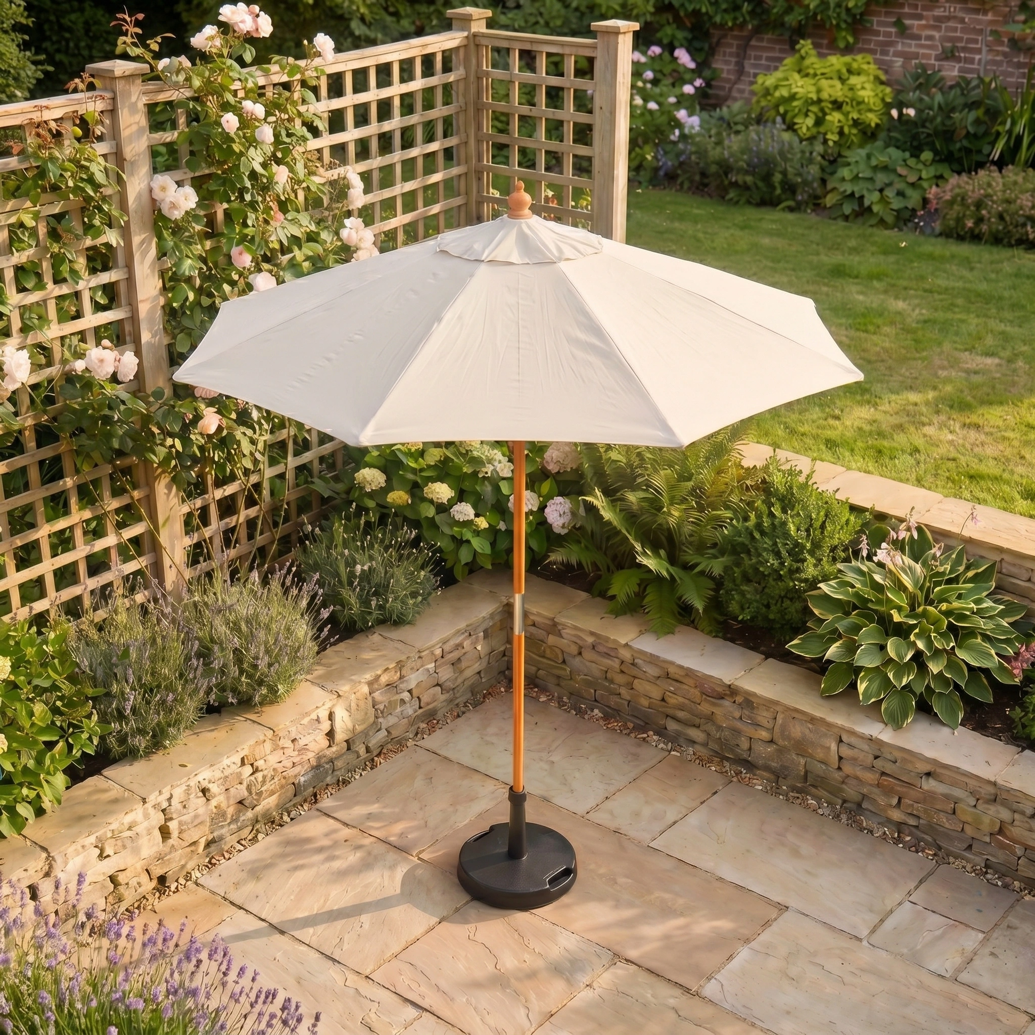 The Kratos 2m Round Wooden Parasol in Beige stands on a stone terrace, surrounded by lush green plants and flowers in a serene garden setting.