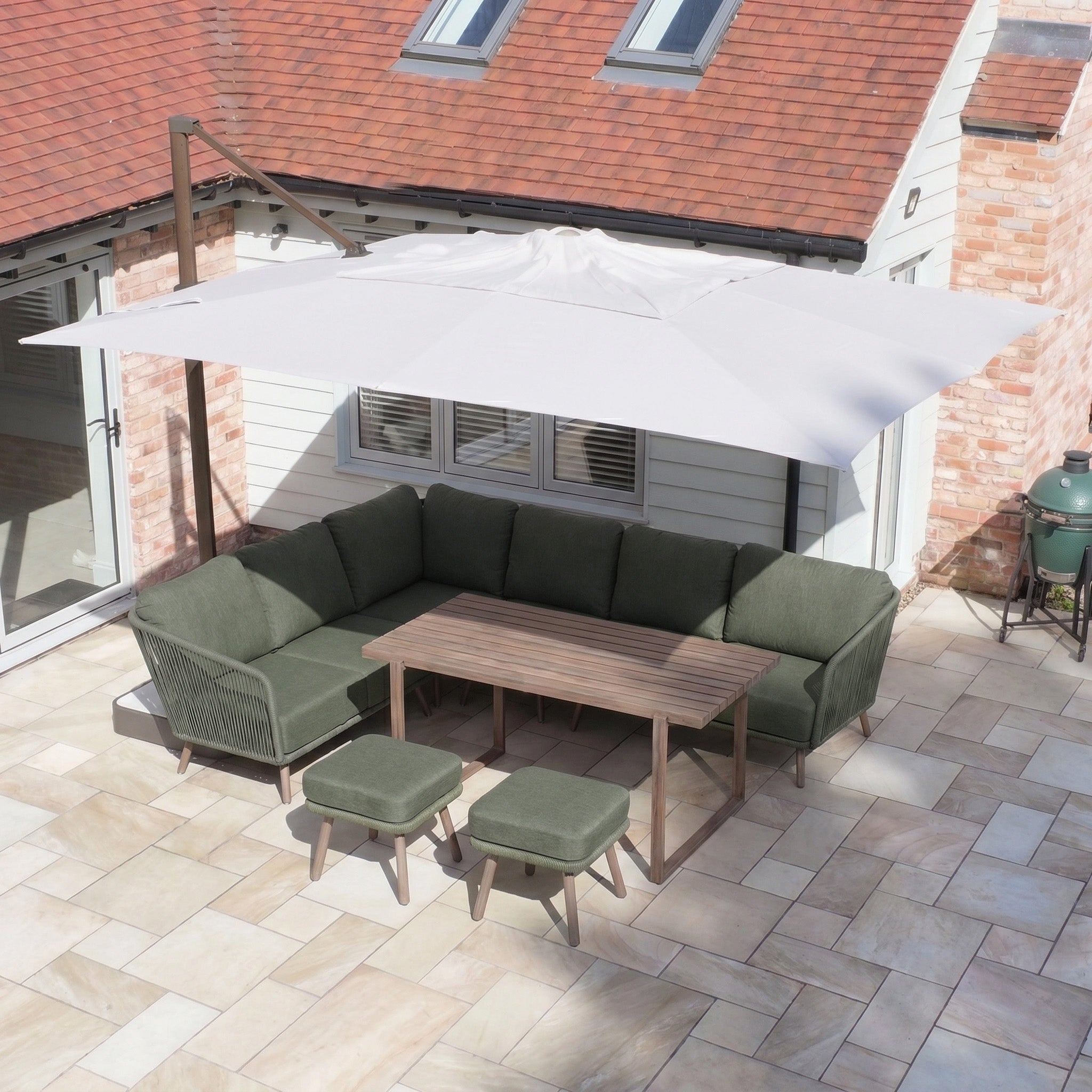 Finca 4m x 3m Cantilever Parasol in Walnut provides sun protection for a patio set with green cushions, a wooden table, and two stools on a tiled patio outside the house.