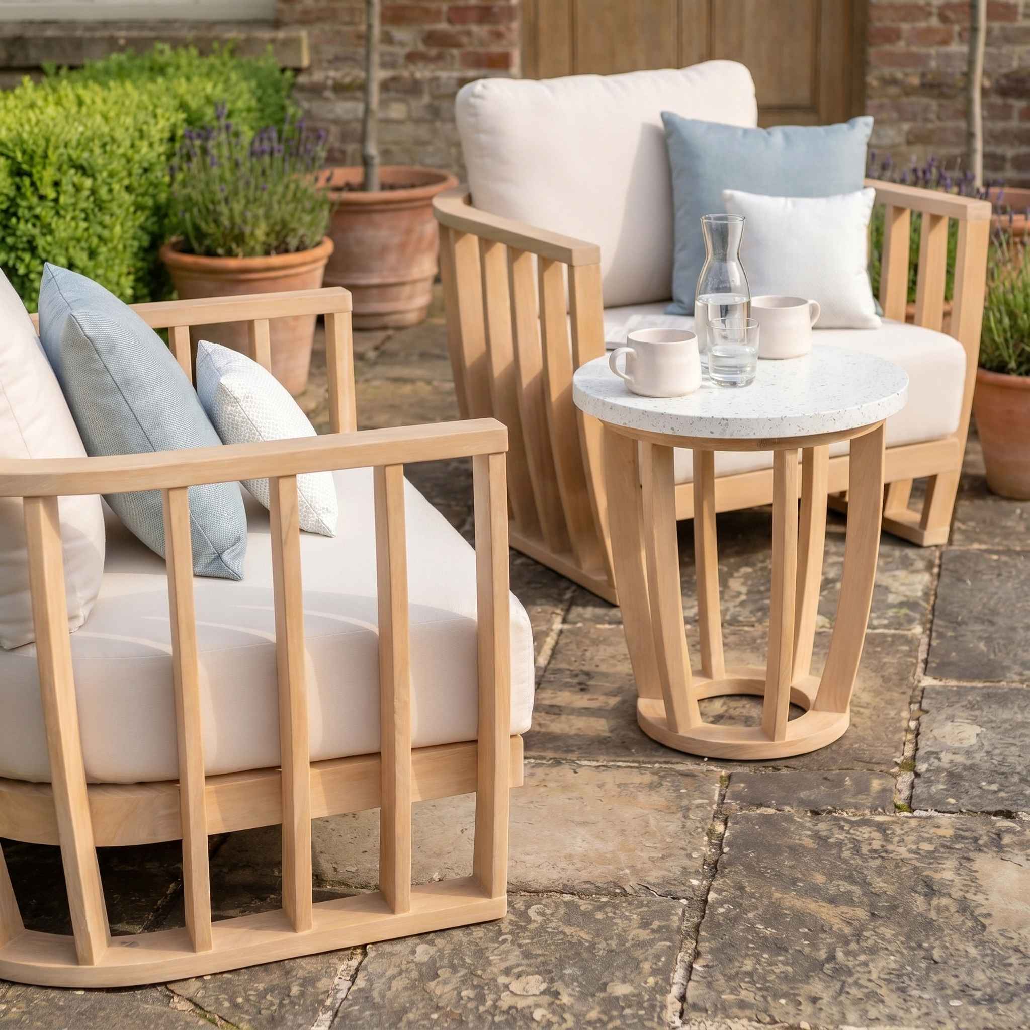 The Cape Lounge Set in Beige, featuring two wooden patio chairs with cushions and a terrazzo coffee table, paired with cups and a glass carafe, creates an inviting outdoor setting.