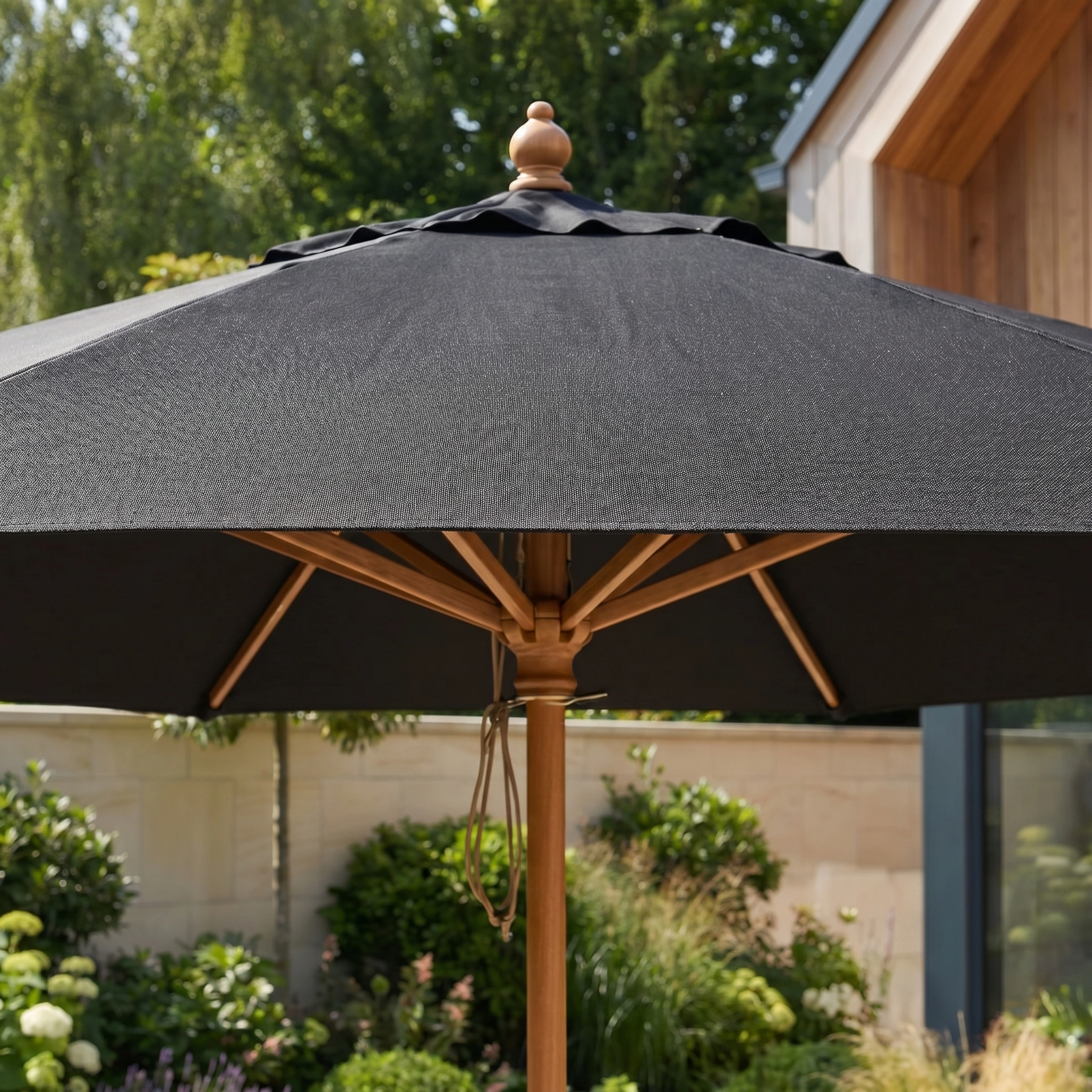 The Kratos 2.5m Round Wooden Parasol in Black stands in a garden, providing UV protection, with lush greenery and a modern house as the backdrop.