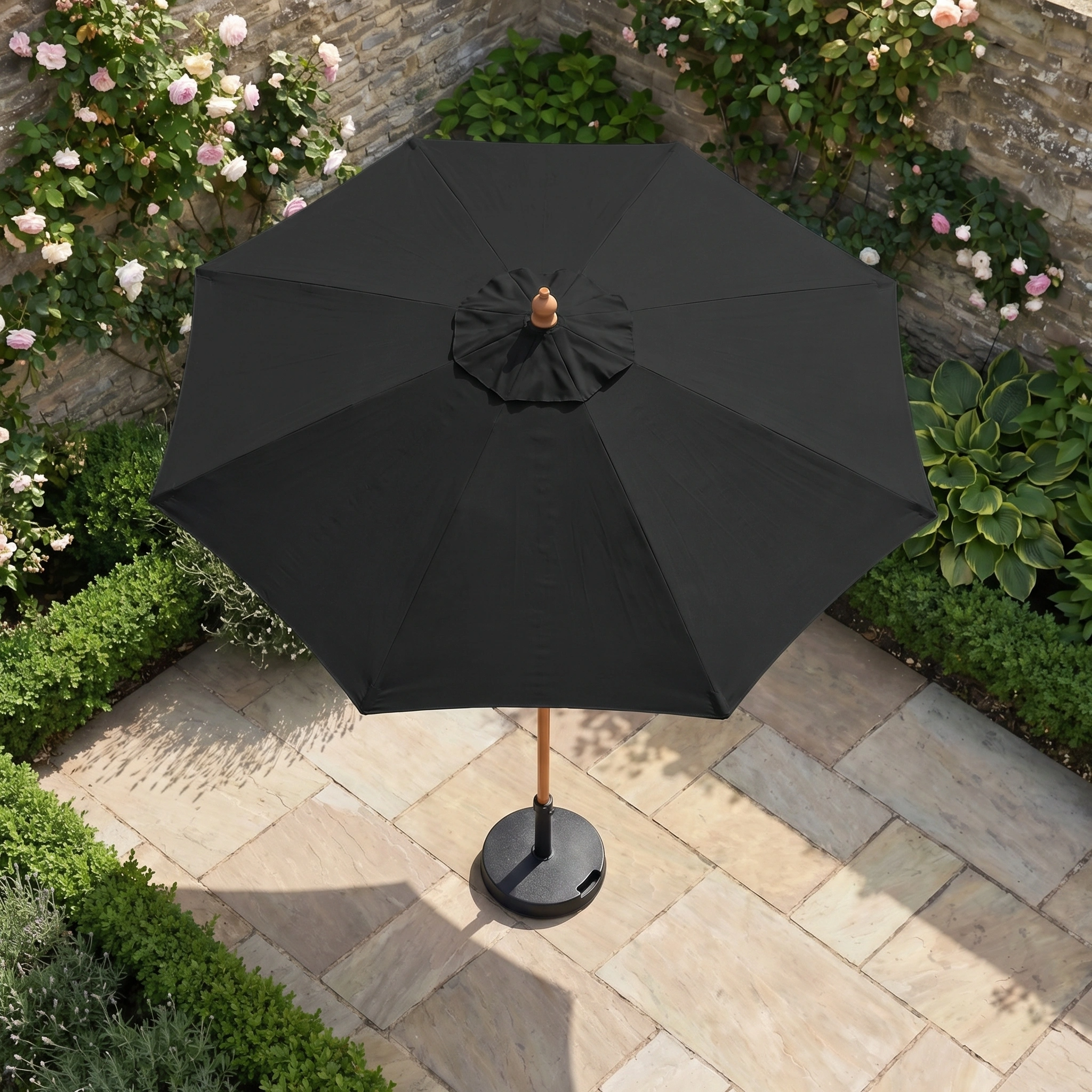 The Kratos 2m Round Wooden Parasol in Black stands on a stone patio among lush bushes and blooming plants, providing excellent sun protection in a vibrant garden setting.