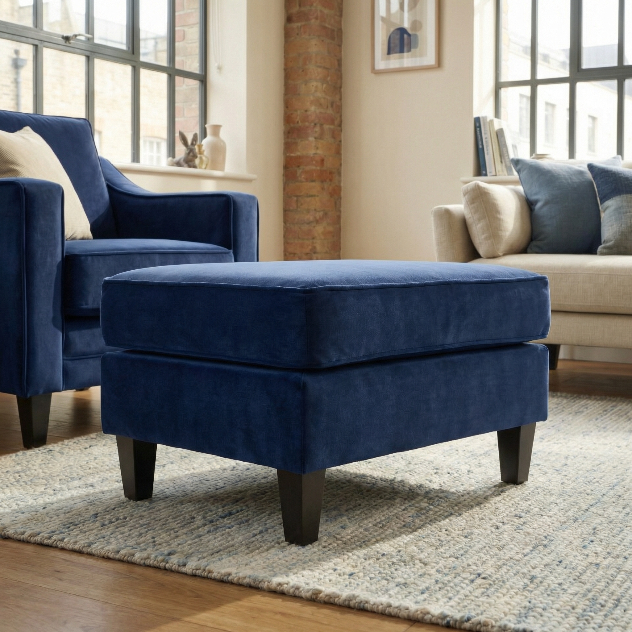 A Wiltshire Velvet Footstool in Royal Blue is placed on a rug in a cozy living room featuring modern furniture and large windows.