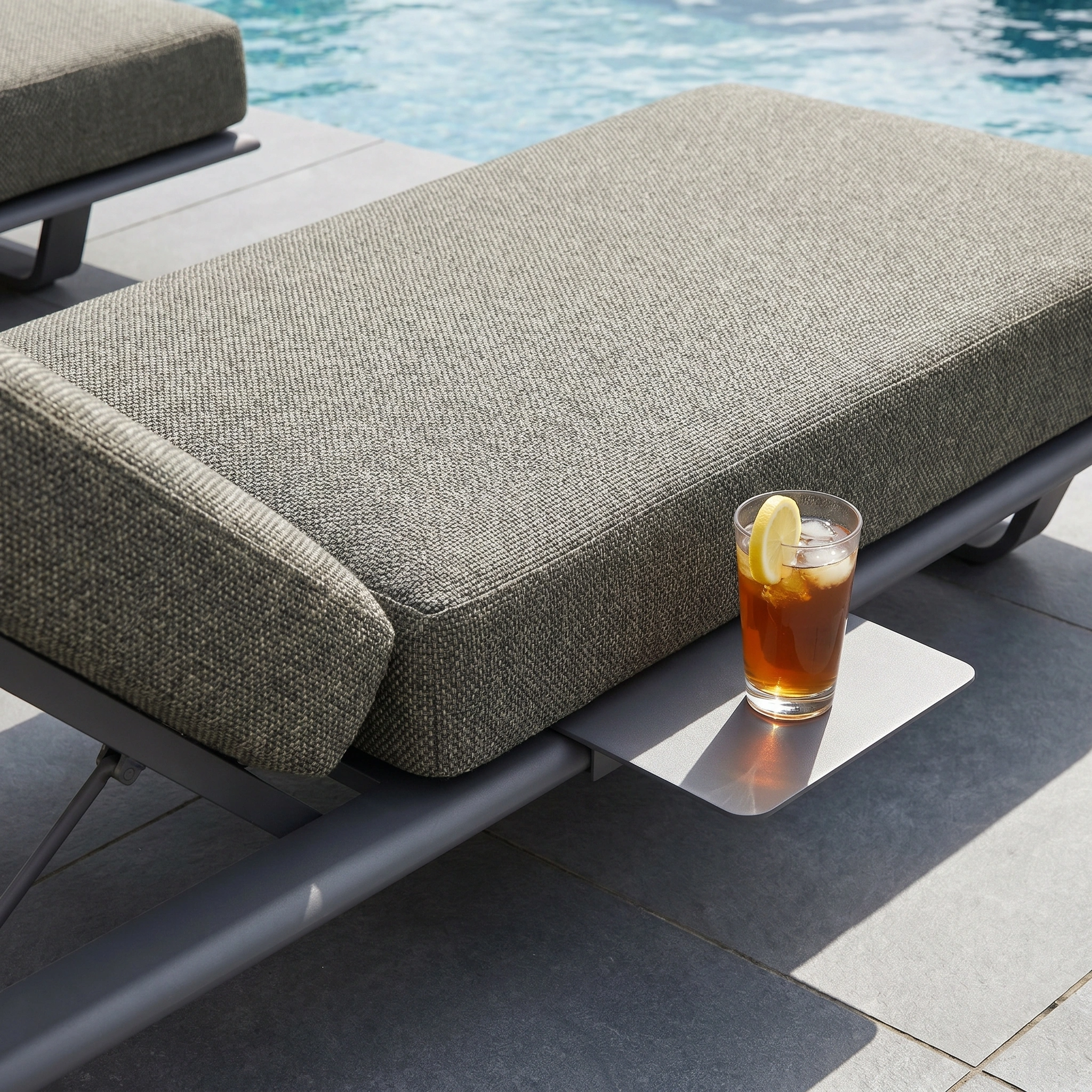 A glass of iced tea with lemon sits on the side table of the Hatia Single Sun Lounger in Charcoal beside the pool.