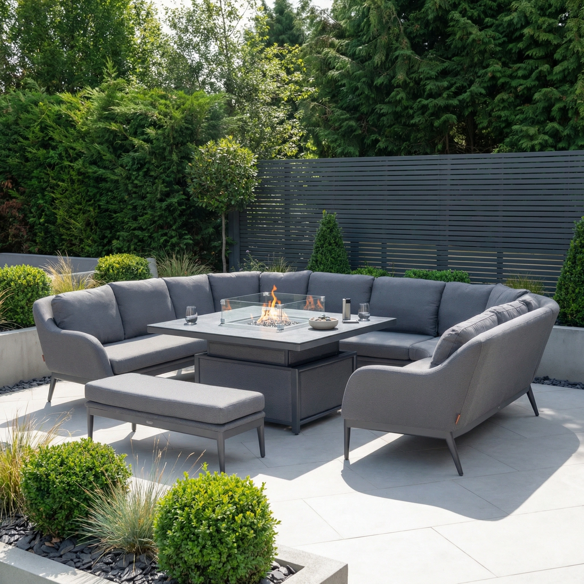 The Luna U-Shape Outdoor Fabric Corner Dining with Rising Firepit Table in Grey features a spacious layout, comfortable bench seating, and a modern rising firepit table, perfect for stylish outdoor dining among lush greenery.