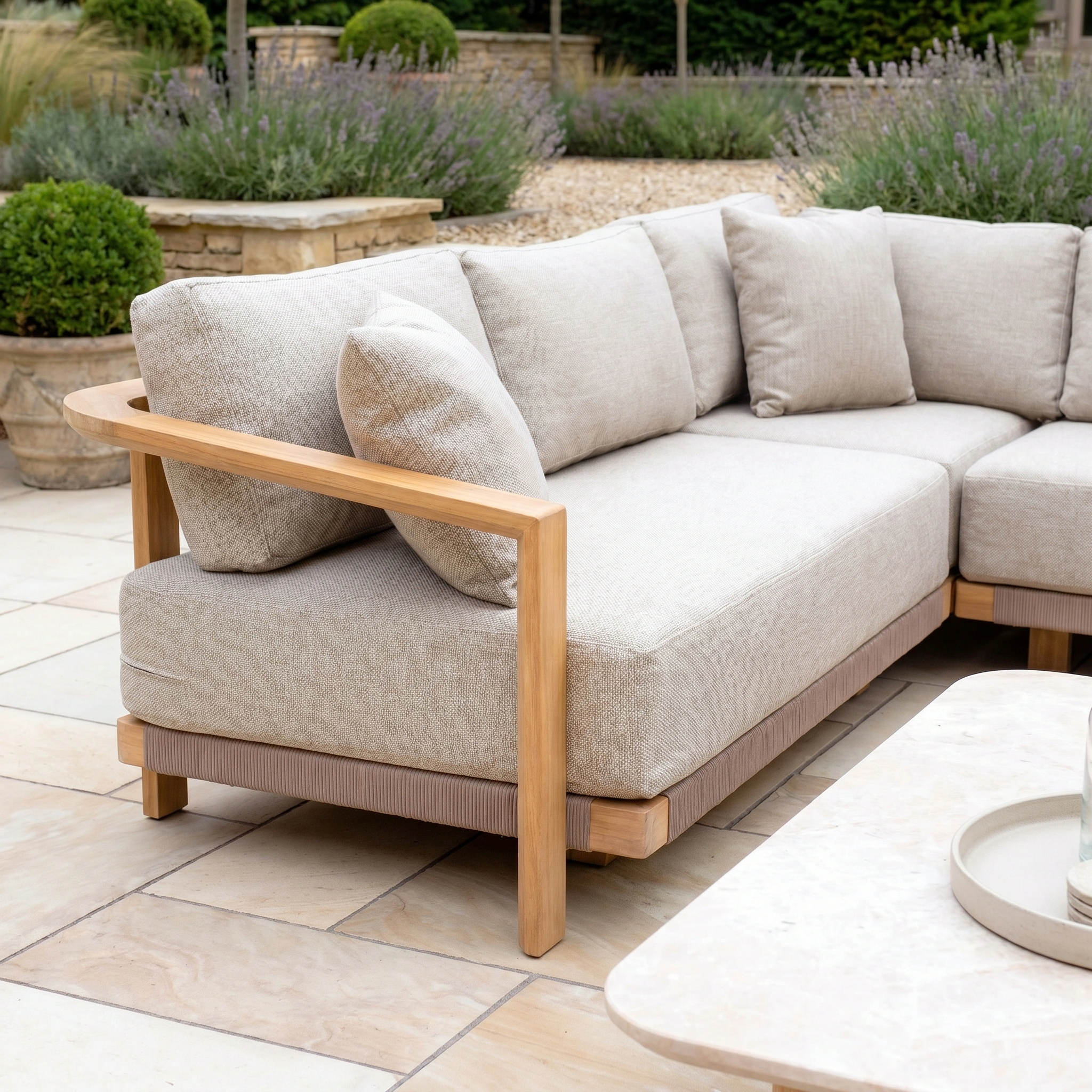 The Tivoli Corner Set with Marble Coffee Table in Fawn features a beige outdoor sectional and light wood frame on a stone patio, surrounded by greenery and plants.