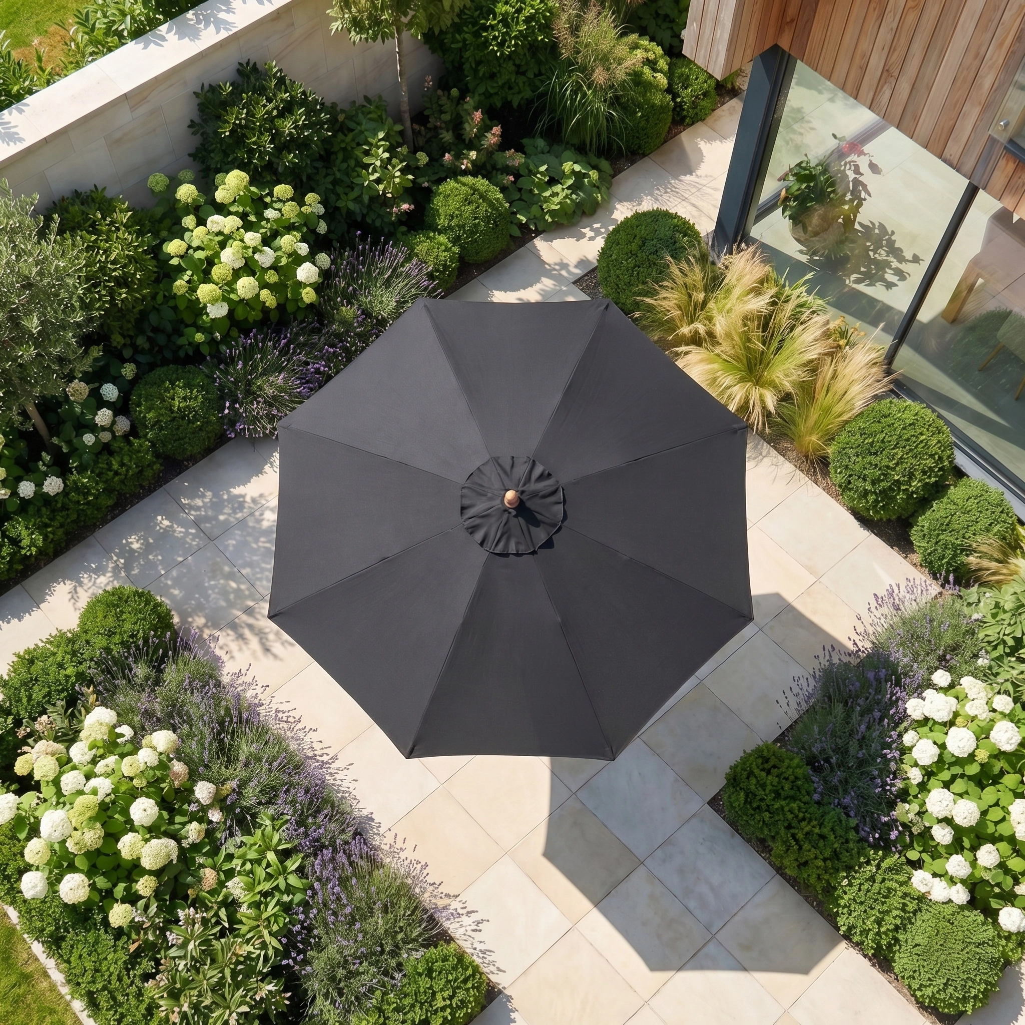 The Kratos 2.5m Round Wooden Parasol in Black with UV protection provides shade for a tiled garden patio surrounded by lush green plants and flowers.