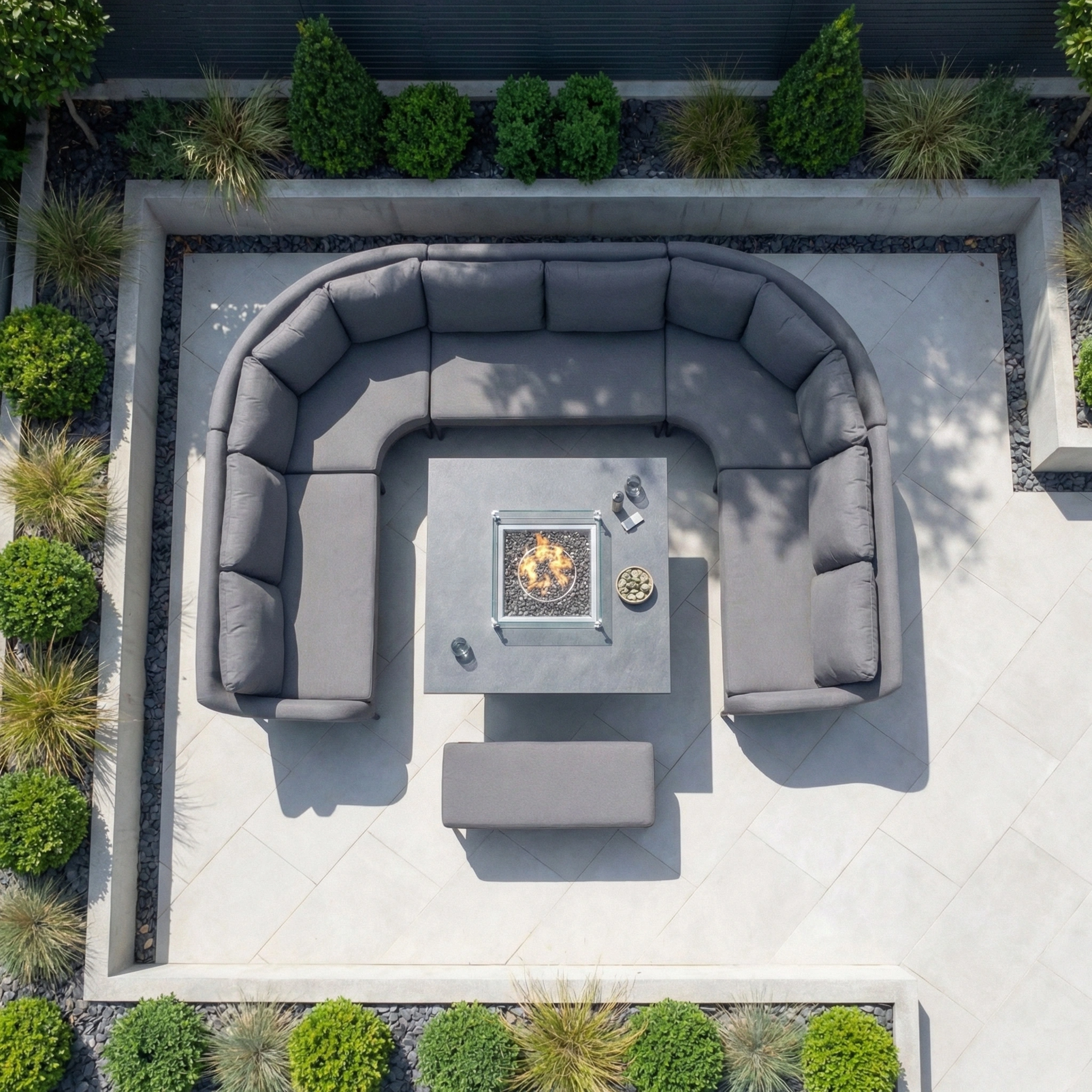The Luna U-Shape Outdoor Fabric Corner Dining with Rising Firepit Table in Grey is set outdoors amid green shrubs and decorative grasses.