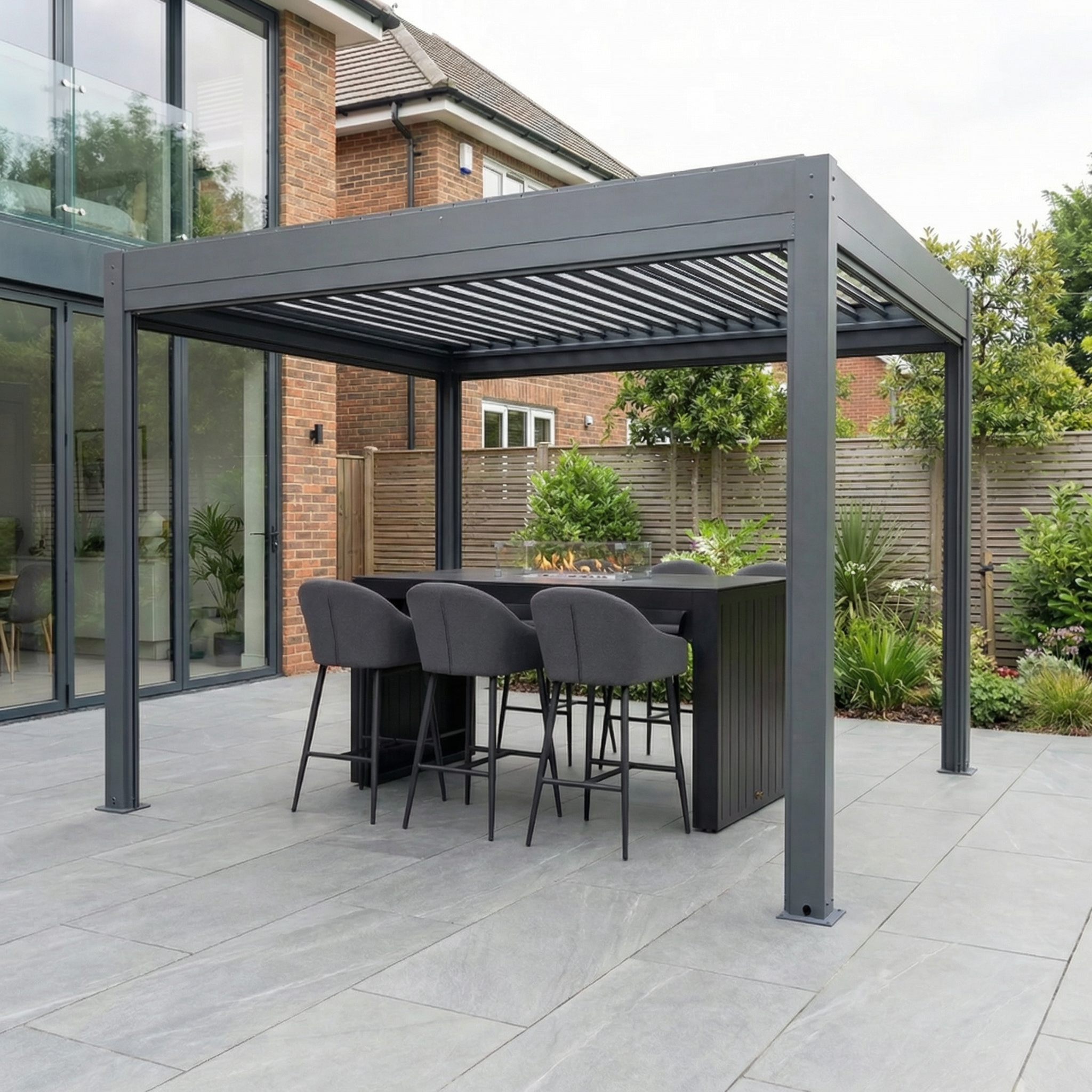 A modern patio showcases the PergoSTET Plus 3m x 3m Square Pergola with manual side blinds and LED lighting in grey, accompanied by a bar table and four gray chairs on a tiled surface near the house.
