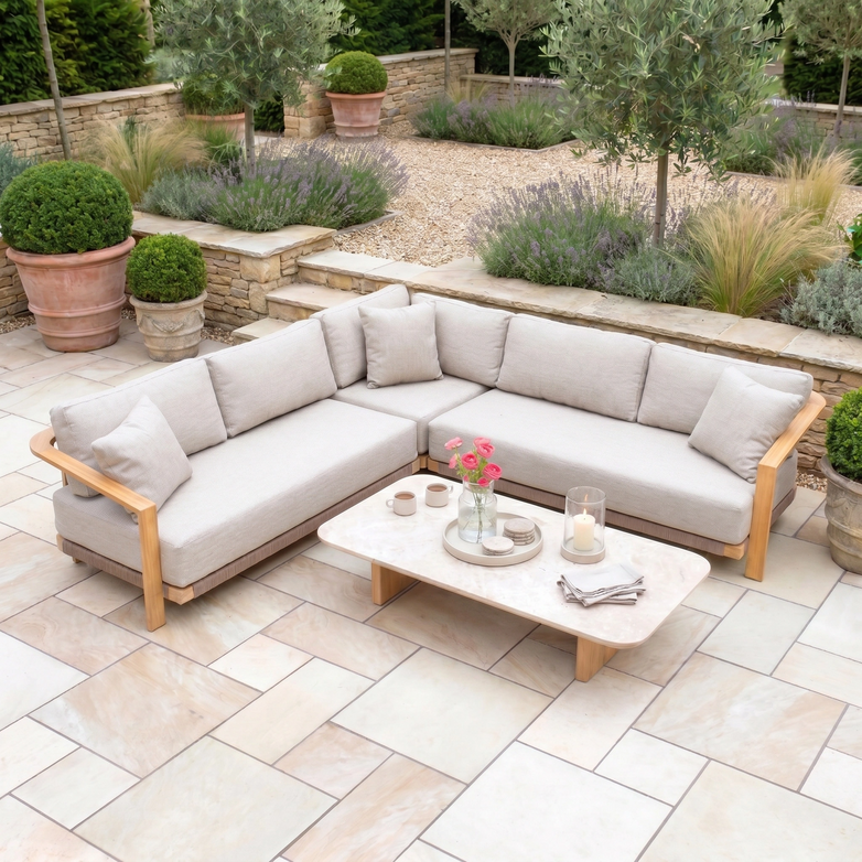 The Tivoli Corner Set with Marble Coffee Table in Fawn sits on a stone patio, accented by flowers, potted plants, and lush greenery.