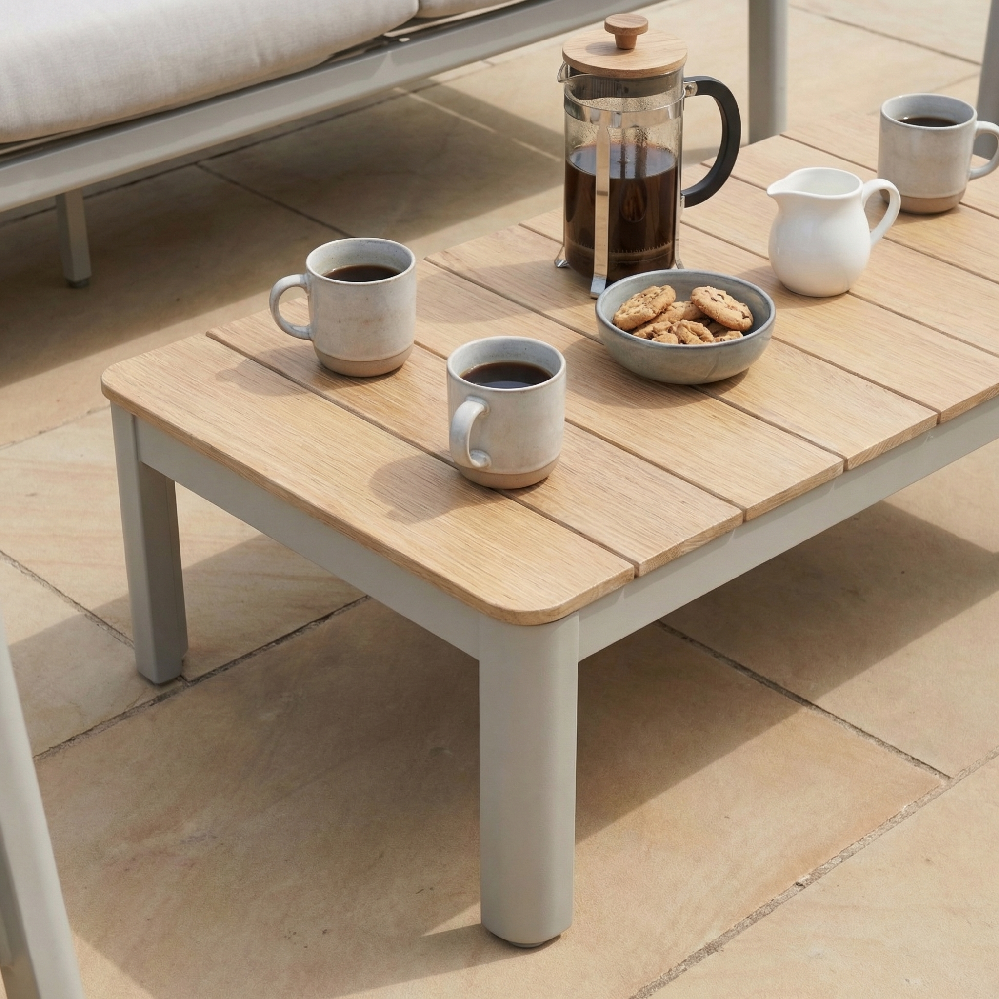 Three coffee mugs, a French press, a bowl of cookies, and a milk jug rest on a wooden table—ideal for sharing treats with friends on your weather-resistant outdoor furniture or the Cora 3 Seat Sofa Set in Latte.