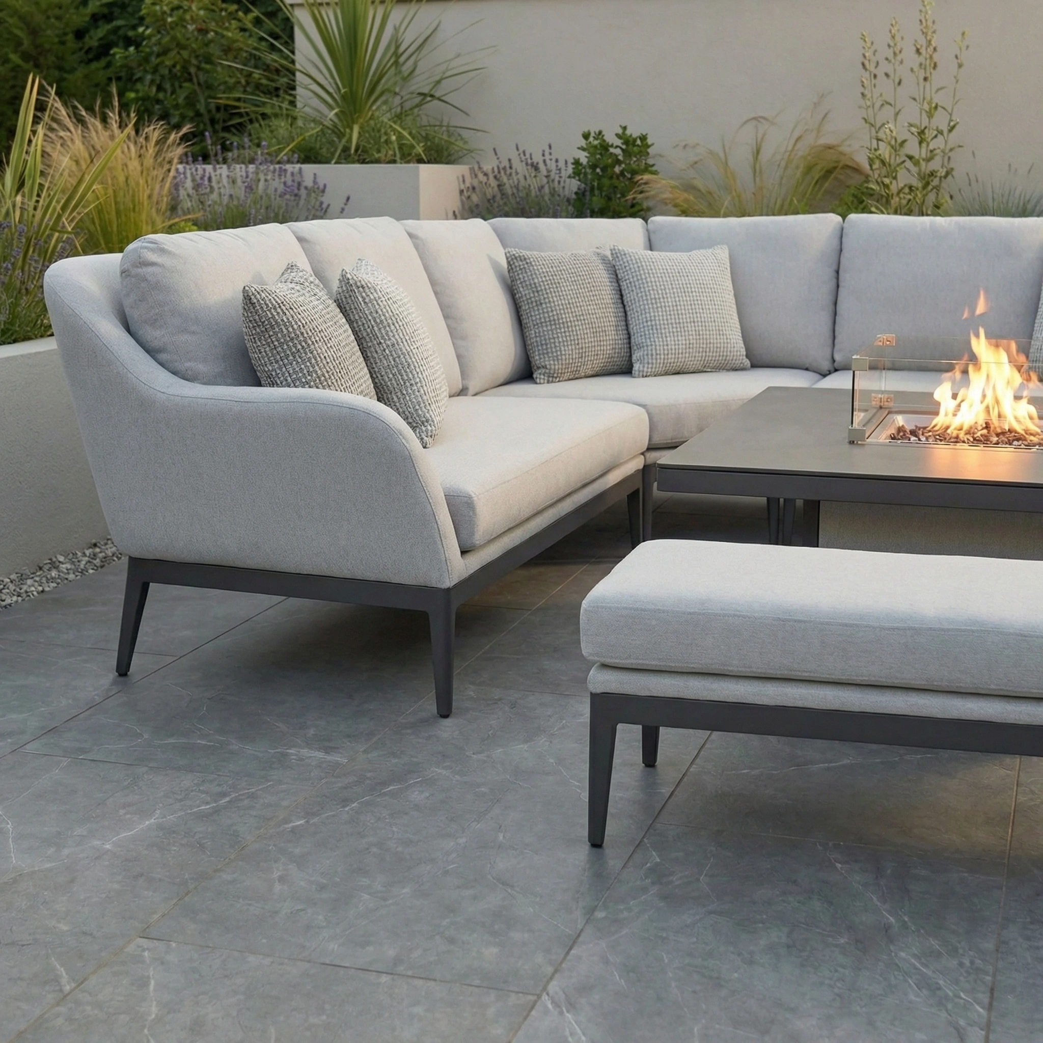 Modern patio with the Luna Deluxe Outdoor Fabric Square Corner Dining Set in Oyster Grey, featuring a rising firepit table and lush green plants in the background.