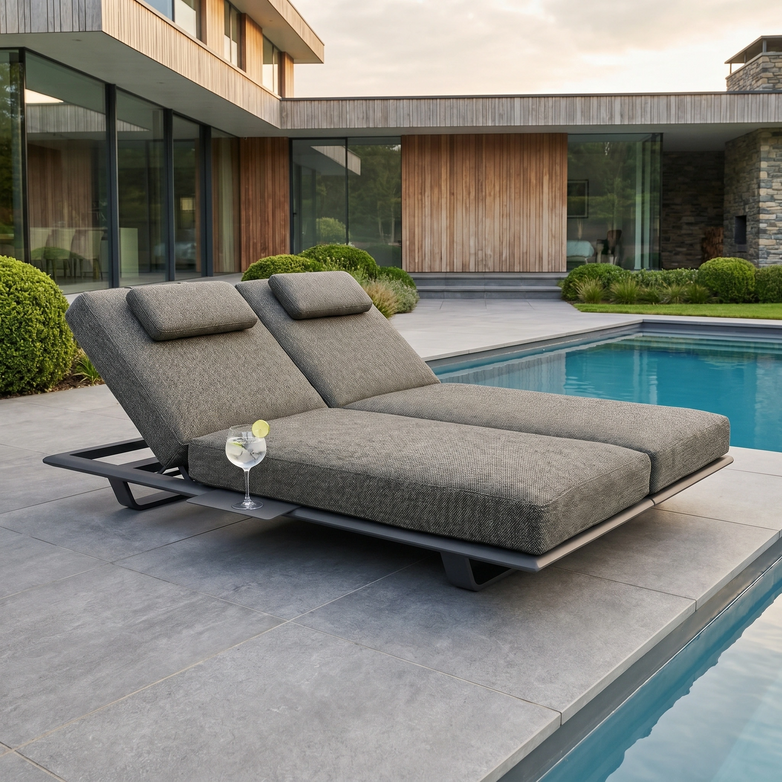 The Hatia Double Sun Lounger with Side Table in Charcoal is set by a modern pool, featuring plush cushions and a drink on the side, perfect for relaxing in a beautifully landscaped backyard.