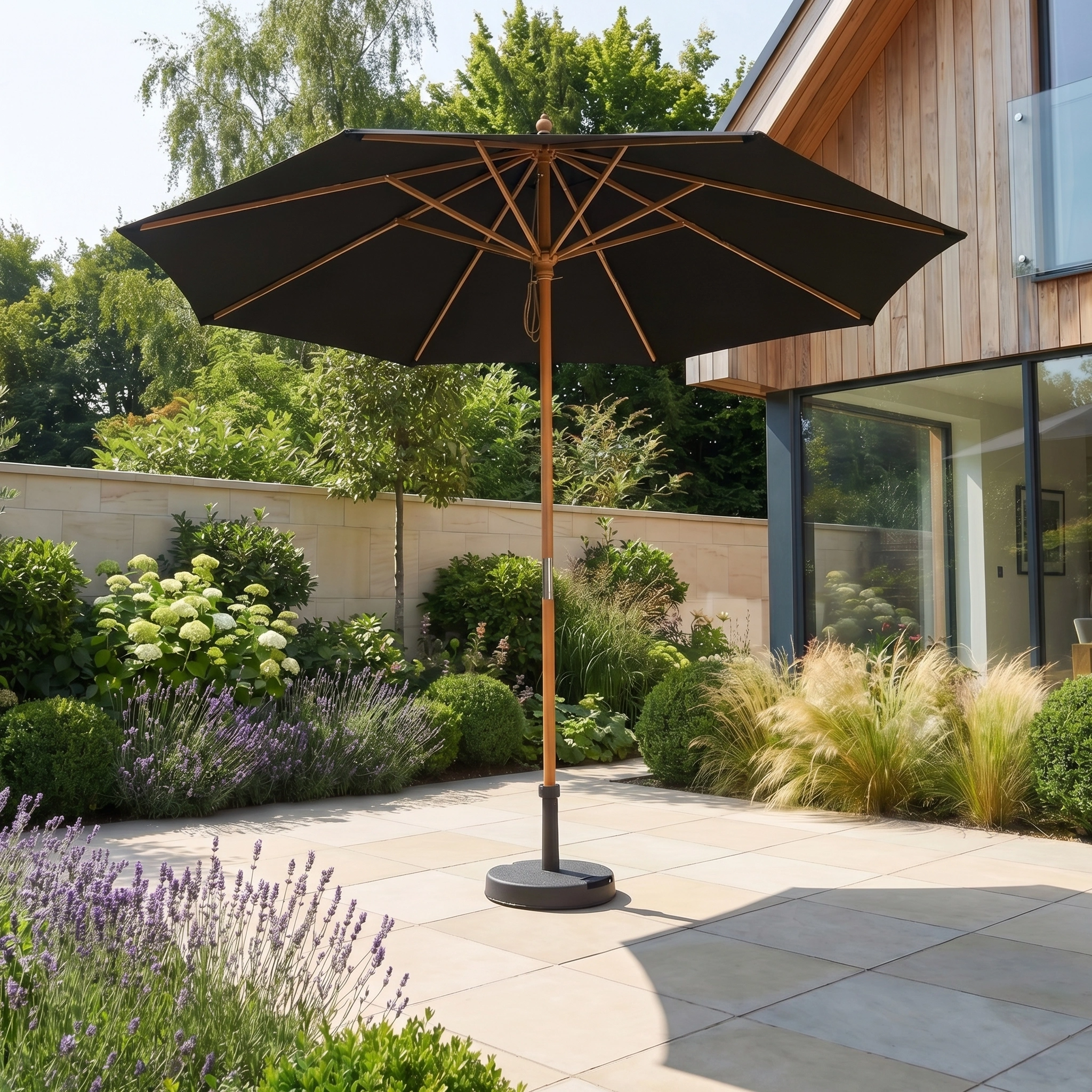 The Kratos 2.5m Round Wooden Parasol in Black stands on a sunny patio amid lush garden plants, offering UV protection with its collapsible design.