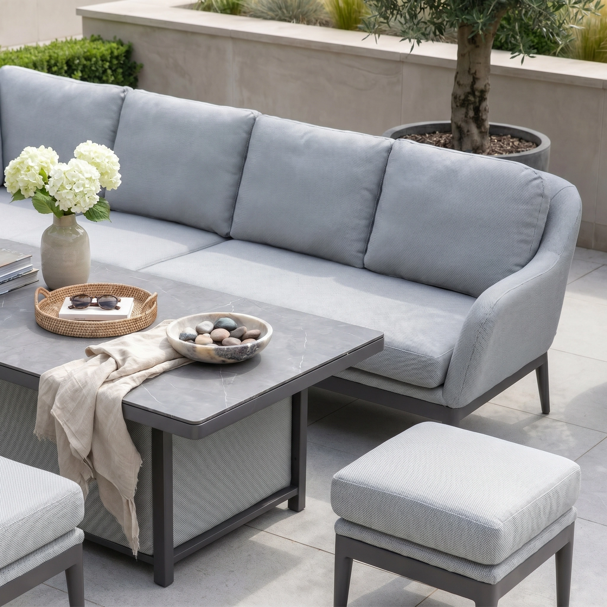 Modern outdoor patio with the Luna Outdoor Fabric Rectangular Corner Dining Set (Left Hand) in Oyster Grey, featuring maintenance-free furniture, a light gray sofa, ottoman, rising table with marble top, and white floral decor.