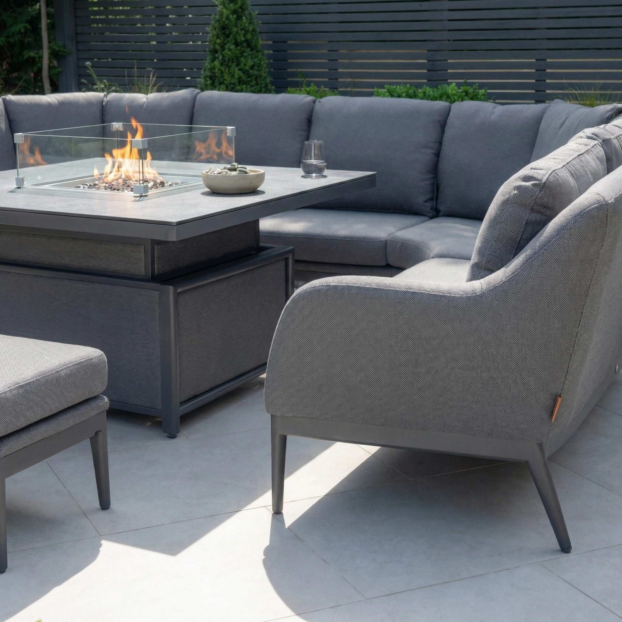 The Luna U-Shape Outdoor Fabric Corner Dining with Rising Firepit Table in Grey sits on patio tiles, enclosed by a privacy fence.