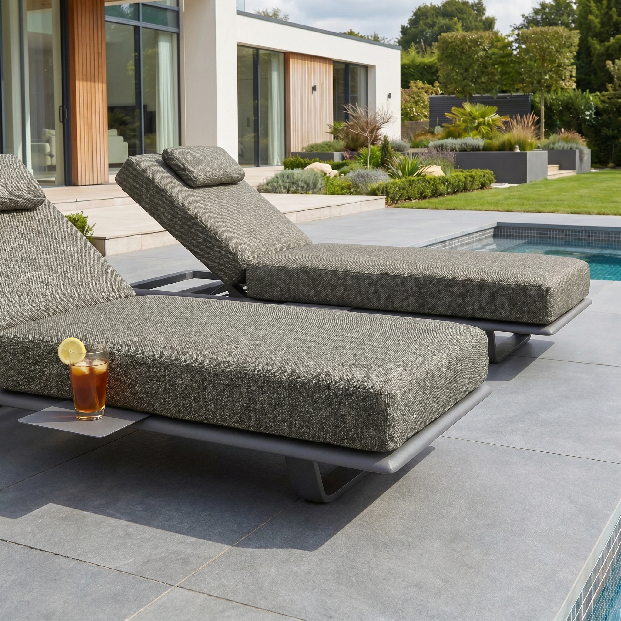 The Hatia Single Sun Lounger with Side Table in Charcoal, made from UV-resistant outdoor material, sits poolside holding iced tea in a landscaped backyard.