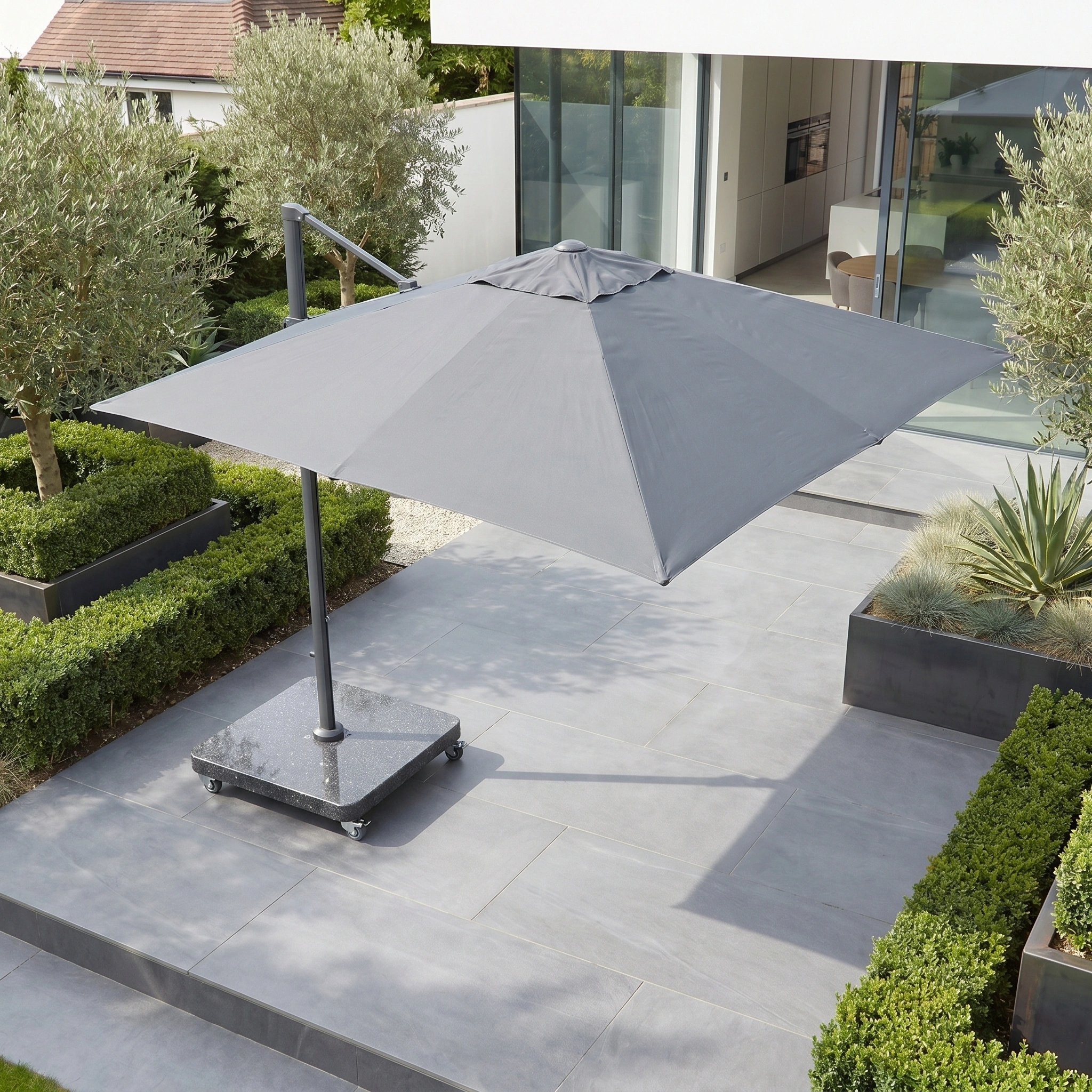 The Glow Challenger T2 3m Square Cantilever Parasol in Anthracite with LED lights stands on a modern stone terrace next to a contemporary house, surrounded by lush green plants and shrubs.