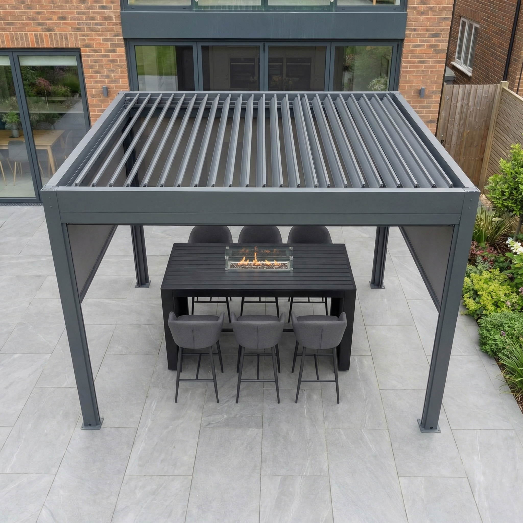The PergoSTET Plus 3m x 3m Square Pergola in grey features 3 manual side blinds, LED lighting, and built-in drainage, providing stylish shelter over a black table with six chairs and a built-in tabletop fire feature.