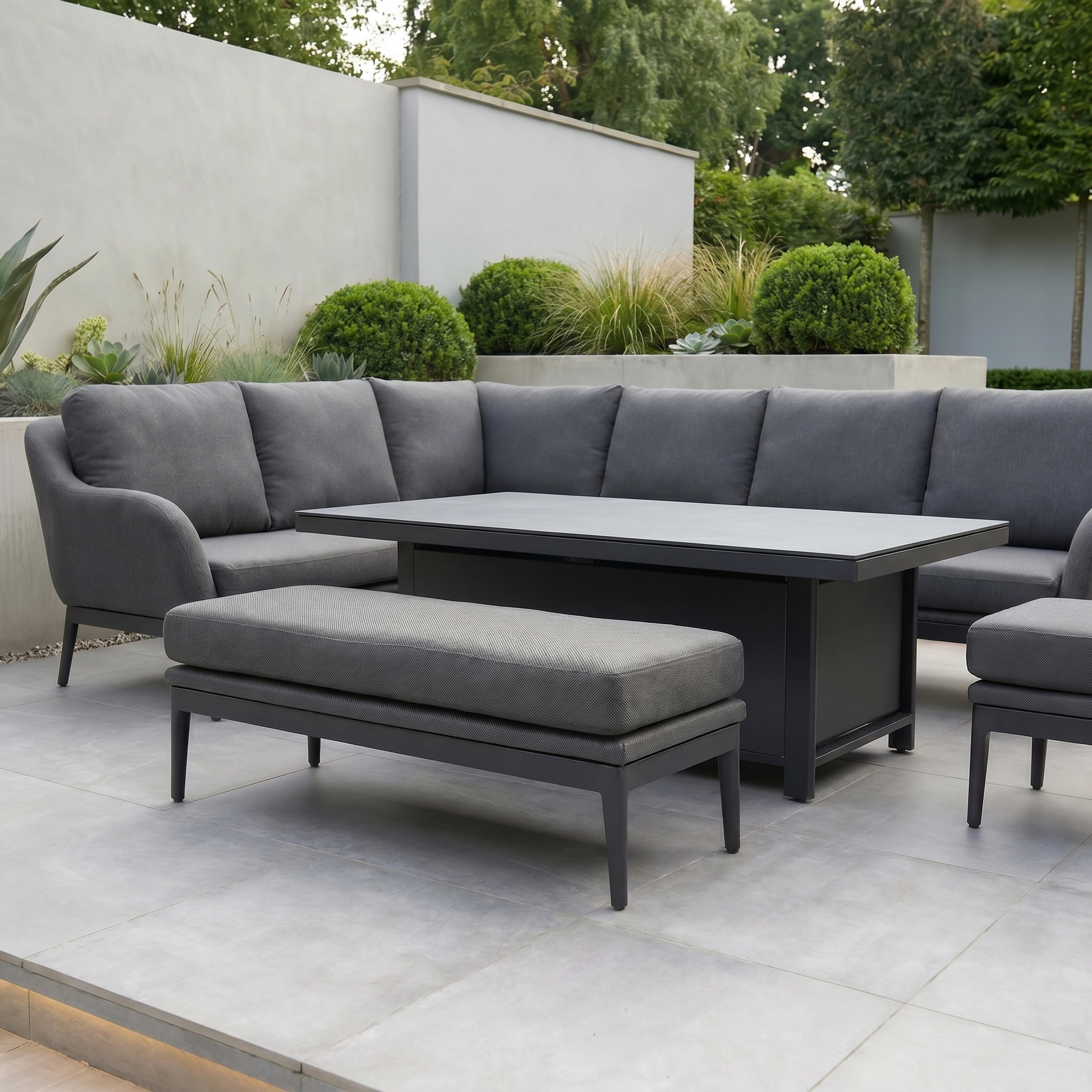 The Luna Outdoor Fabric Rectangular Corner Dining Set with Rising Table in Grey (Left Hand) features gray cushions and is displayed on a tiled patio surrounded by greenery and bushes.