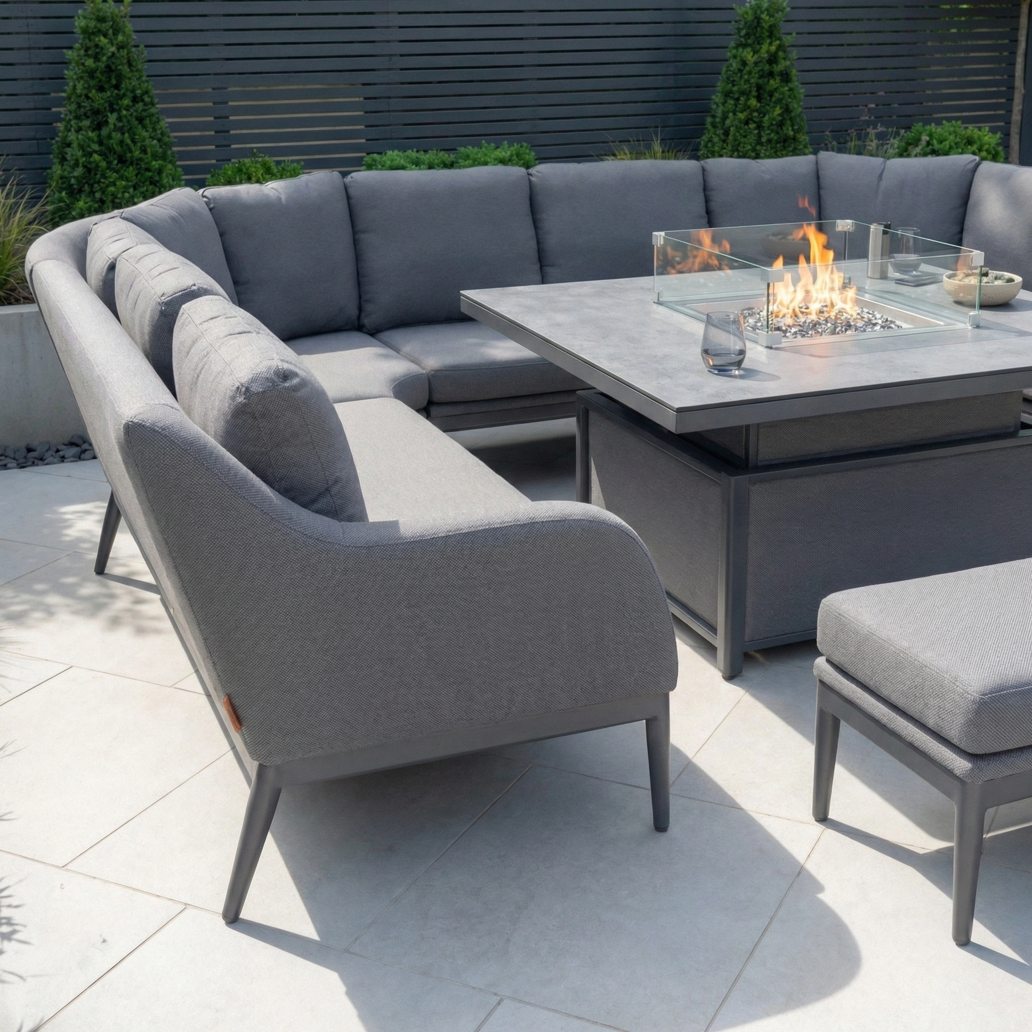 The Luna U-Shape Outdoor Fabric Corner Dining set in grey features a rising firepit table with a glass wind guard, creating an ideal modern setup for outdoor dining and relaxation.