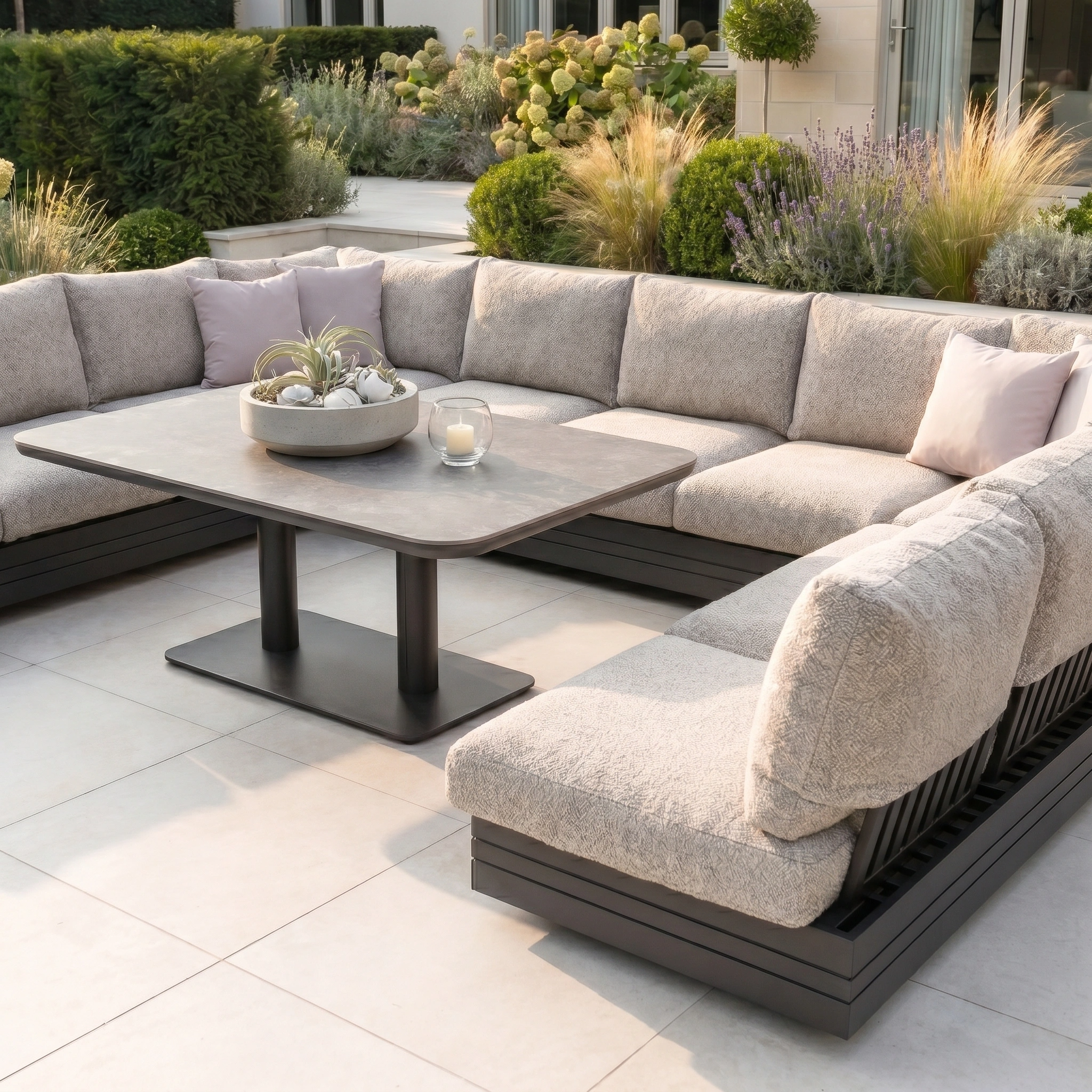 The Savona Luxury Outdoor Large U-Shape Corner Sofa Set with Altura Rising Table in Truffle creates a modern patio setting, paired with lush plants and crafted from rust-proof aluminium for style and durability.