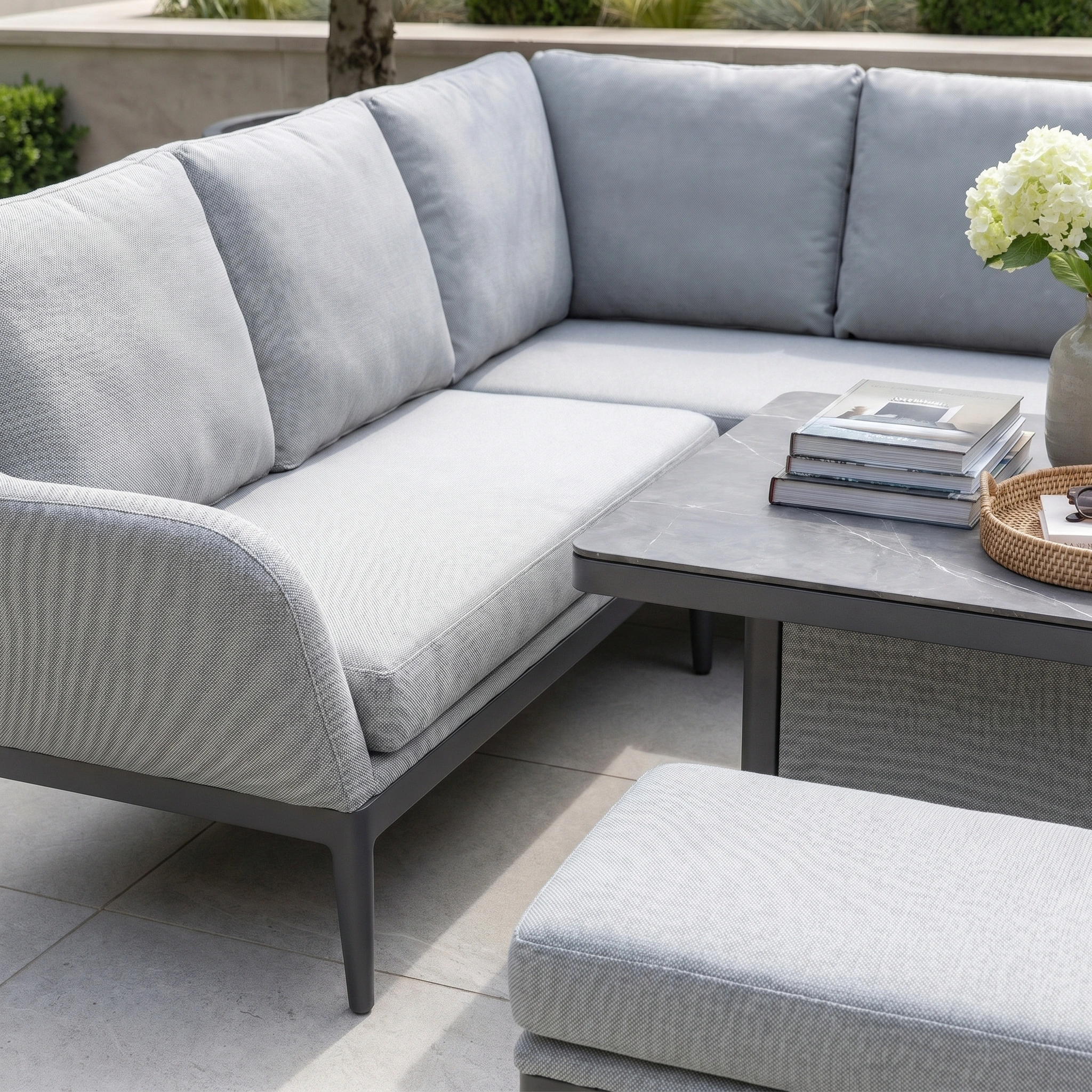 The Luna Outdoor Fabric Rectangular Corner Dining Set with Rising Table in Oyster Grey (Left Hand) offers inviting, maintenance-free garden comfort with modern style, shown here accessorized with books, a tray, and a vase of flowers on the table.