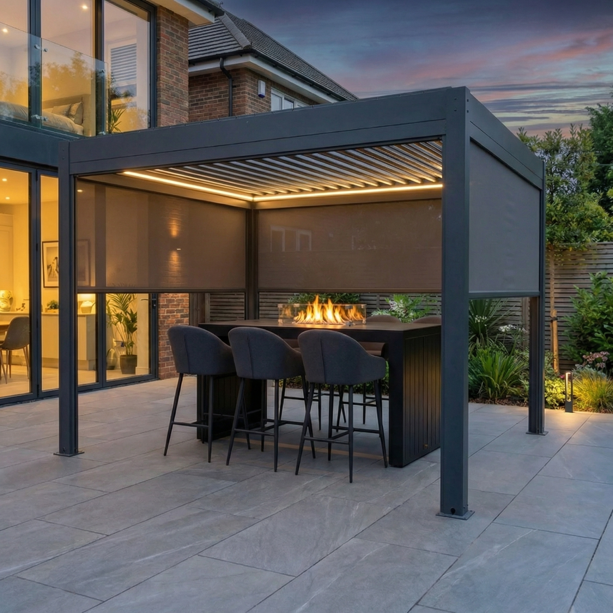 Modern outdoor patio at dusk with a covered seating area, bar stools, fire feature, and the PergoSTET Plus 3m x 3m Square Pergola in grey featuring tilting louvres, manual side blinds, and LED lighting for adjustable shade and style.