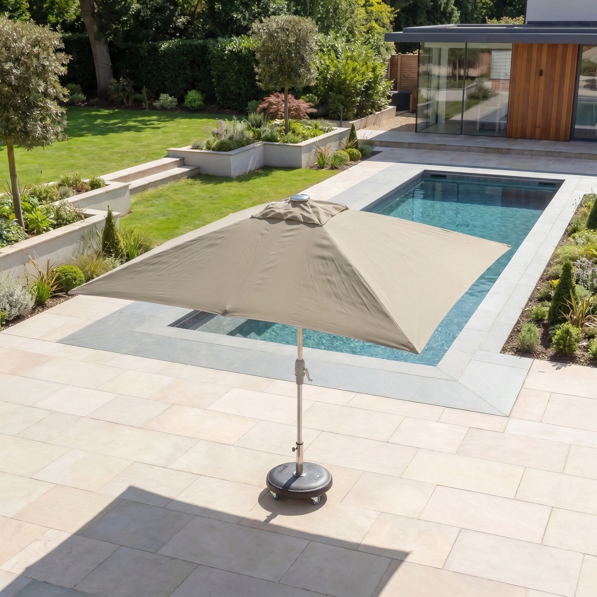 A Zelos 3m x 2m Rectangular Parasol with LED Lighting in taupe offers UV protection beside a modern pool in a landscaped backyard surrounded by trees and greenery.