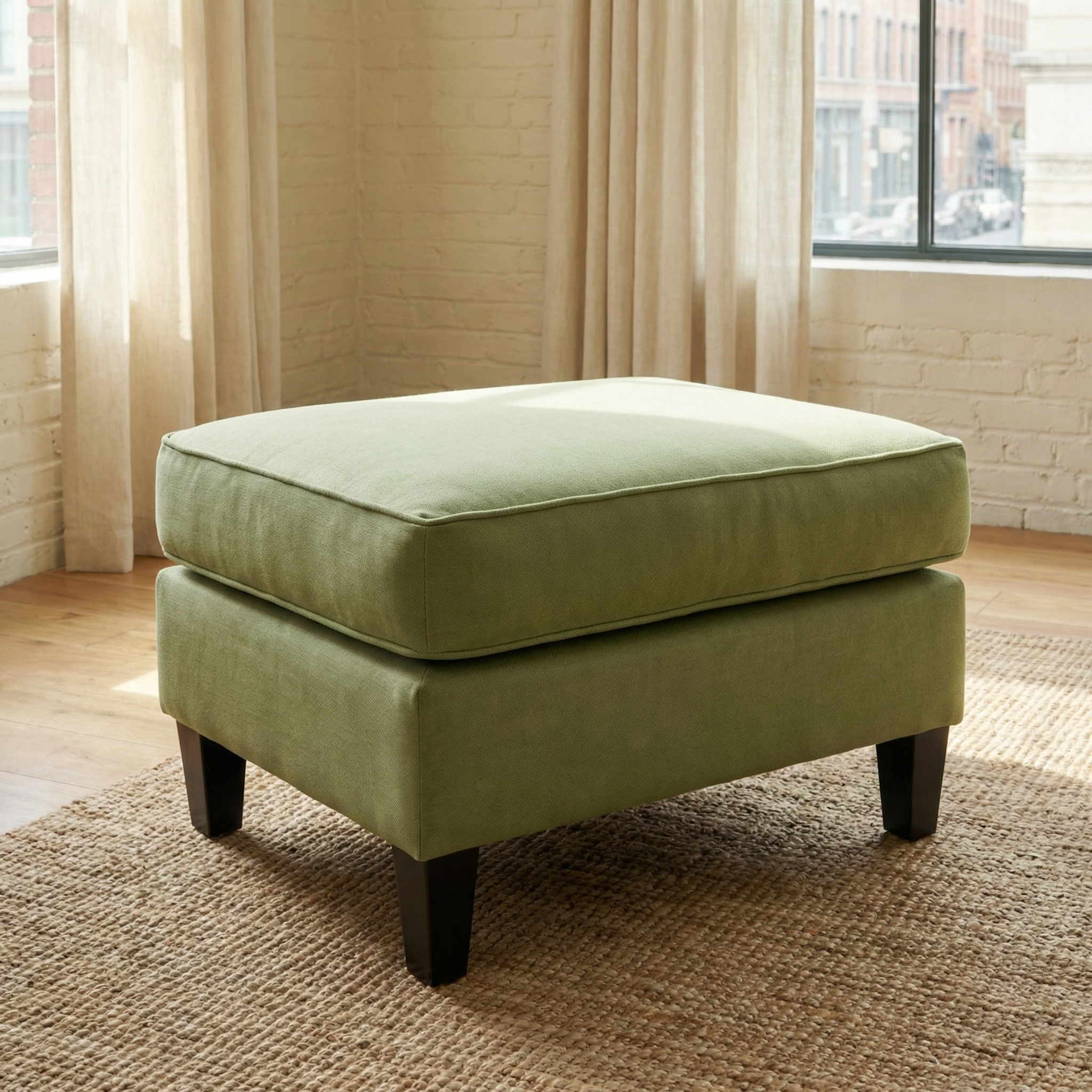 The Wiltshire Footstool in Olive with dark wooden legs sits on a woven rug in a bright room featuring large windows and flowing curtains.