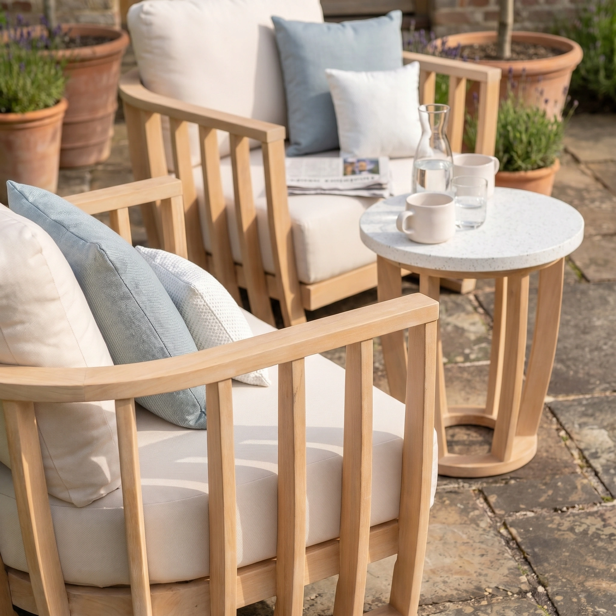The Cape Lounge Set in Beige features two wooden patio chairs with cushions and pillows beside a small terrazzo coffee table, complete with mugs and a glass carafe.
