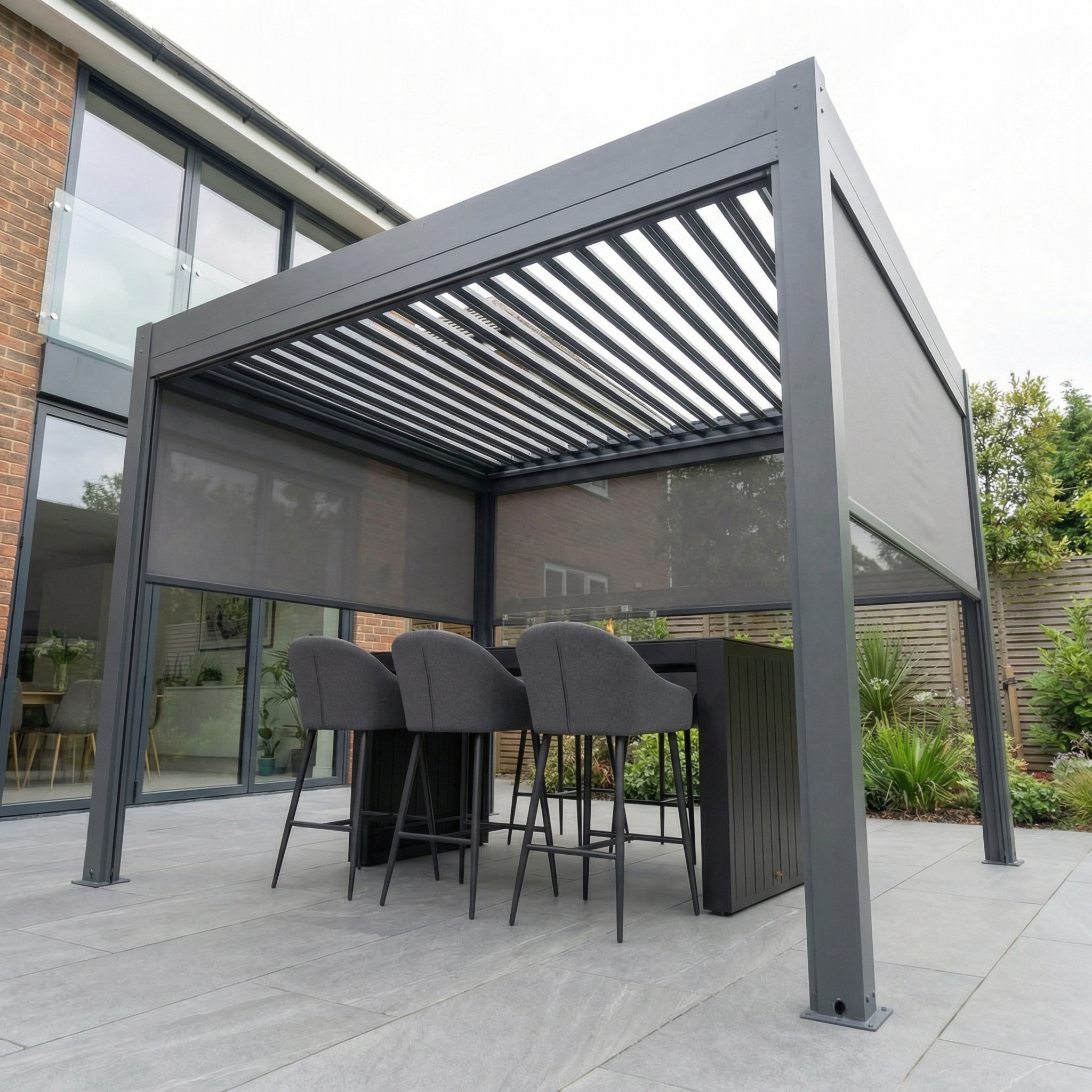PergoSTET Plus 3m x 3m Square Pergola with manual side blinds and LED lighting in grey, shown on a patio beside the house, features a modern design ideal for outdoor relaxation.