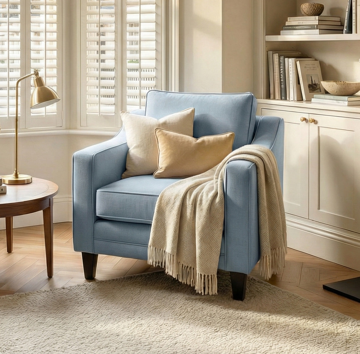 A cozy living room corner with a Wiltshire Armchair in Sky Blue handmade in England, soft cushions, a throw blanket, and bookshelves by a window.
