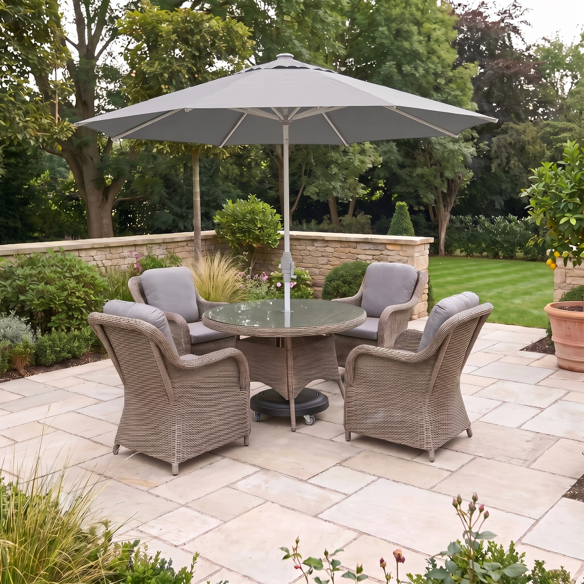 The Isla 4 Seat Round Dining Set in Flint features four weather-resistant wicker chairs with cushions arranged around a round glass table and umbrella on a stone patio in a garden, creating an inviting outdoor dining space ideal for relaxing or entertaining guests.